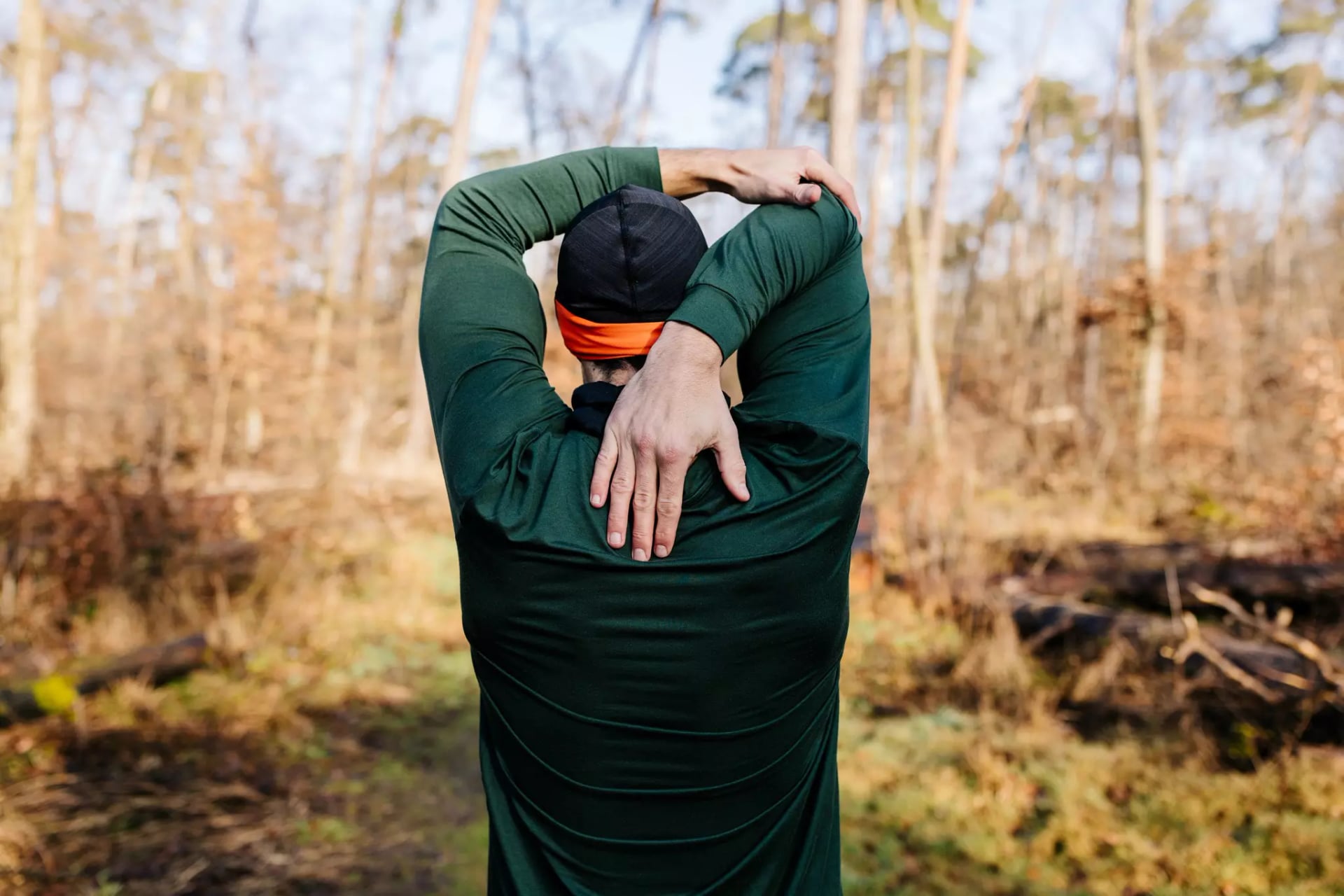 Person von hinten in grünem Langarmshirt und schwarzer Mütze mit orangefarbenem Stirnband, die im Wald den rechten Arm über den Kopf nach links zieht