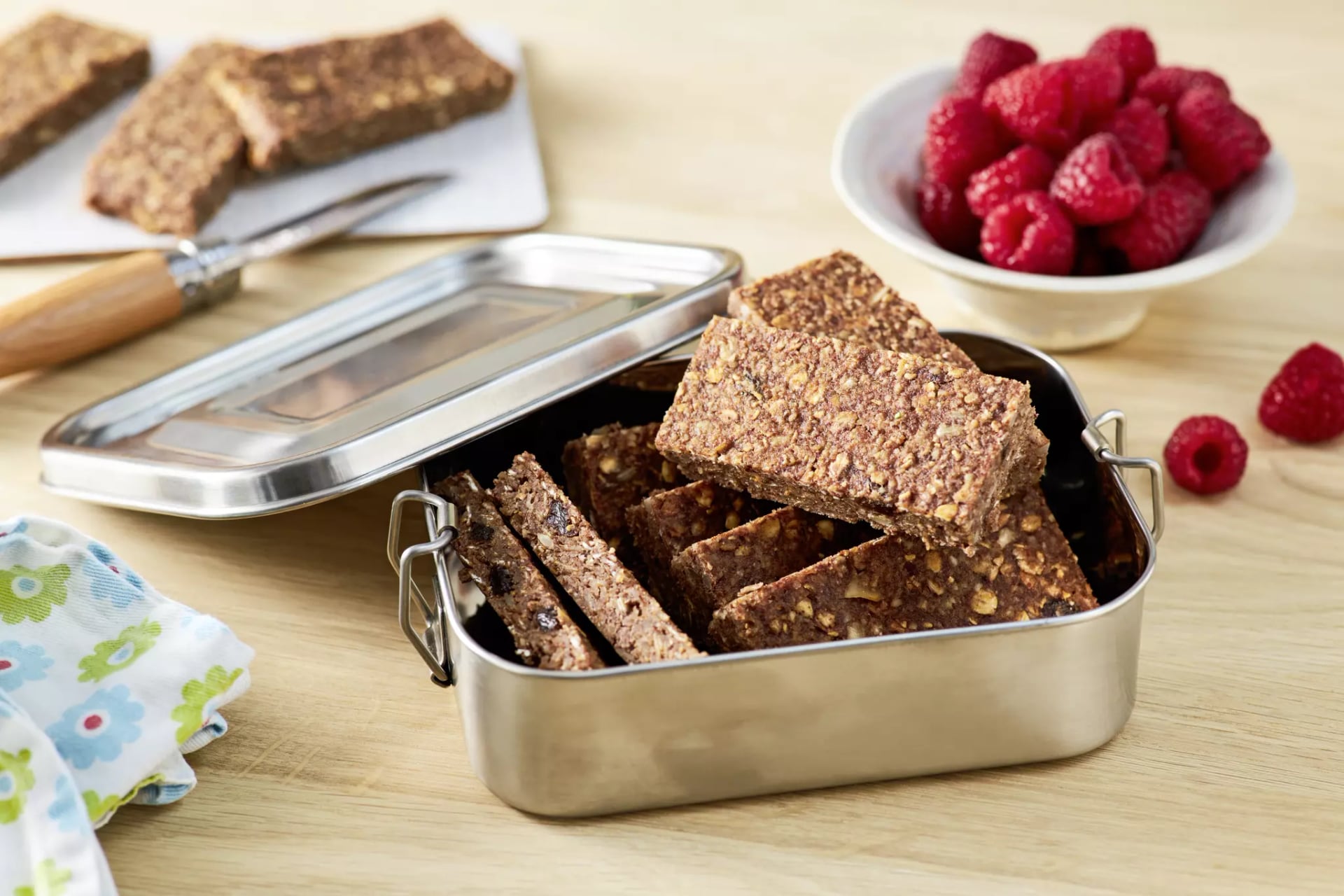 Metallbox mit rechteckigen Müsliriegeln auf einem Holztisch neben einer Schale mit Himbeeren.