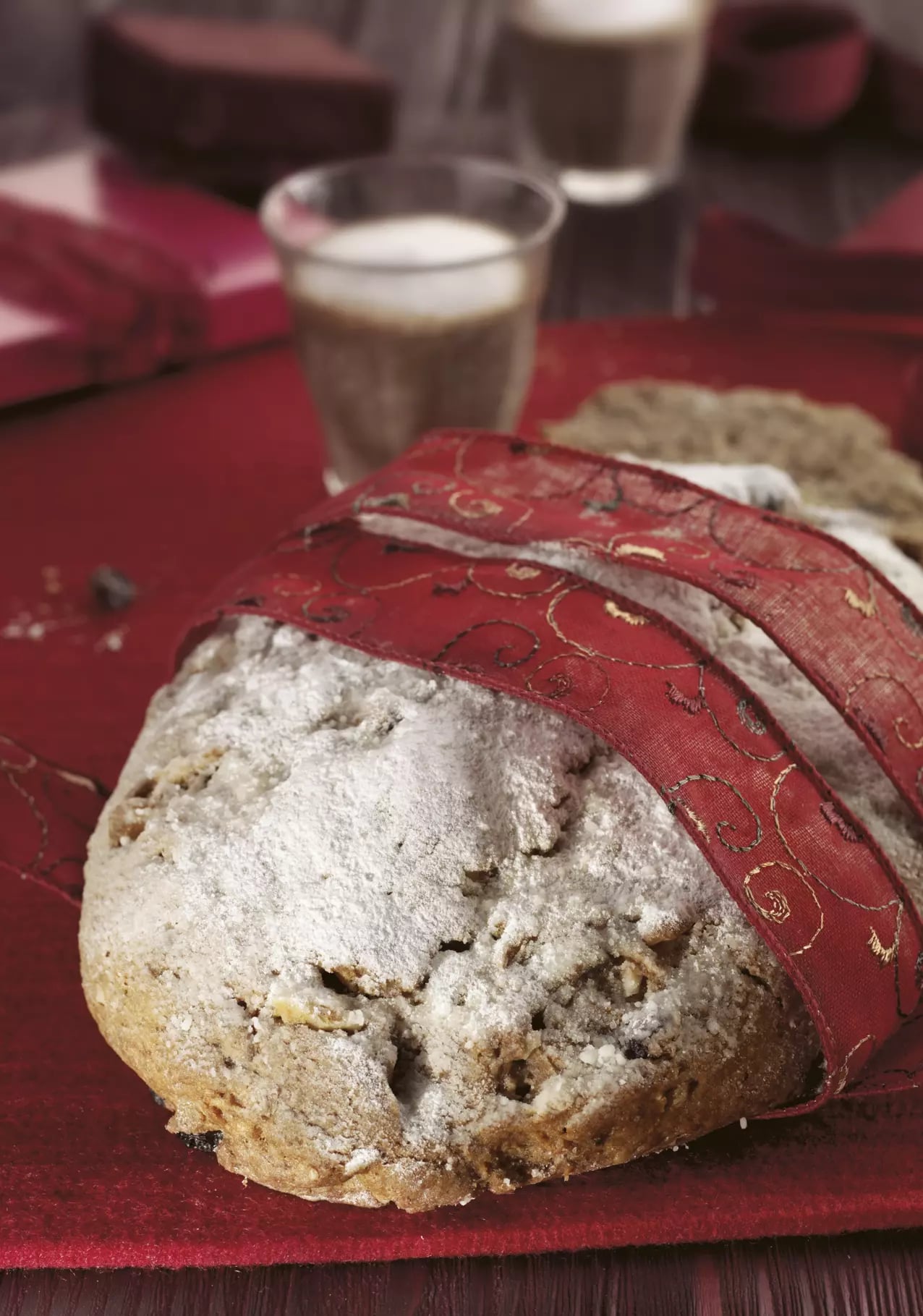 Ganzer Quarkstollen auf einer roten Tischdecke. Der Stollen ist mit einem roten Stoffband umwickelt, welches goldene Details hat.