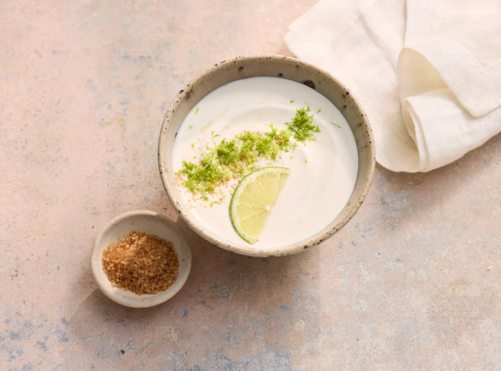 Schale mit Joghurt-Limetten-Dip, garniert mit Limettenscheibe und Limettenabrieb, daneben kleine Schale mit braunem Gewürz und ein Stoffserviettenstück auf hellem Untergrund.