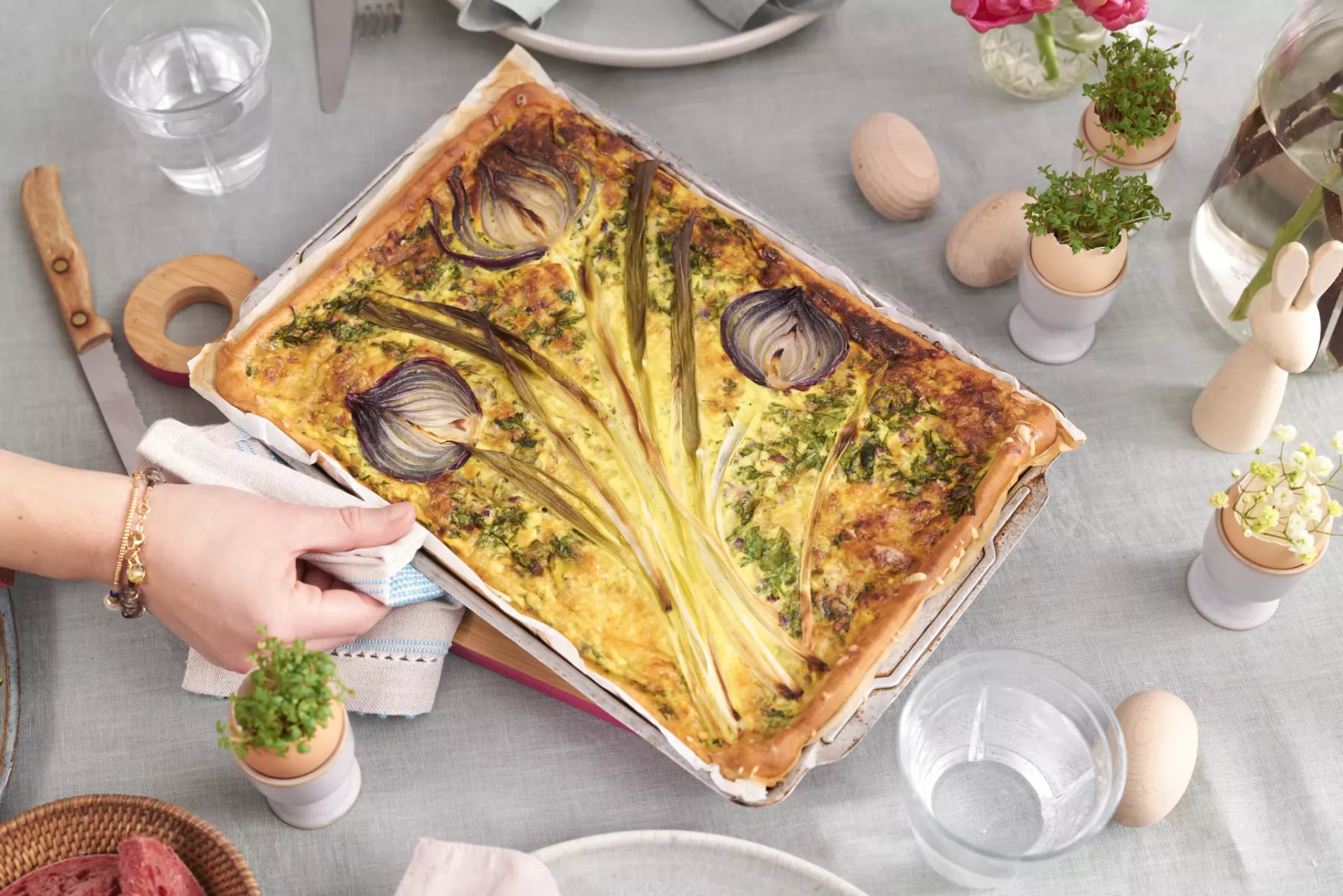 Hand hält rechteckige Gemüsequiche mit Spargel und roten Zwiebeln in Backform auf gedecktem Tisch mit Eiern und Kräutern.