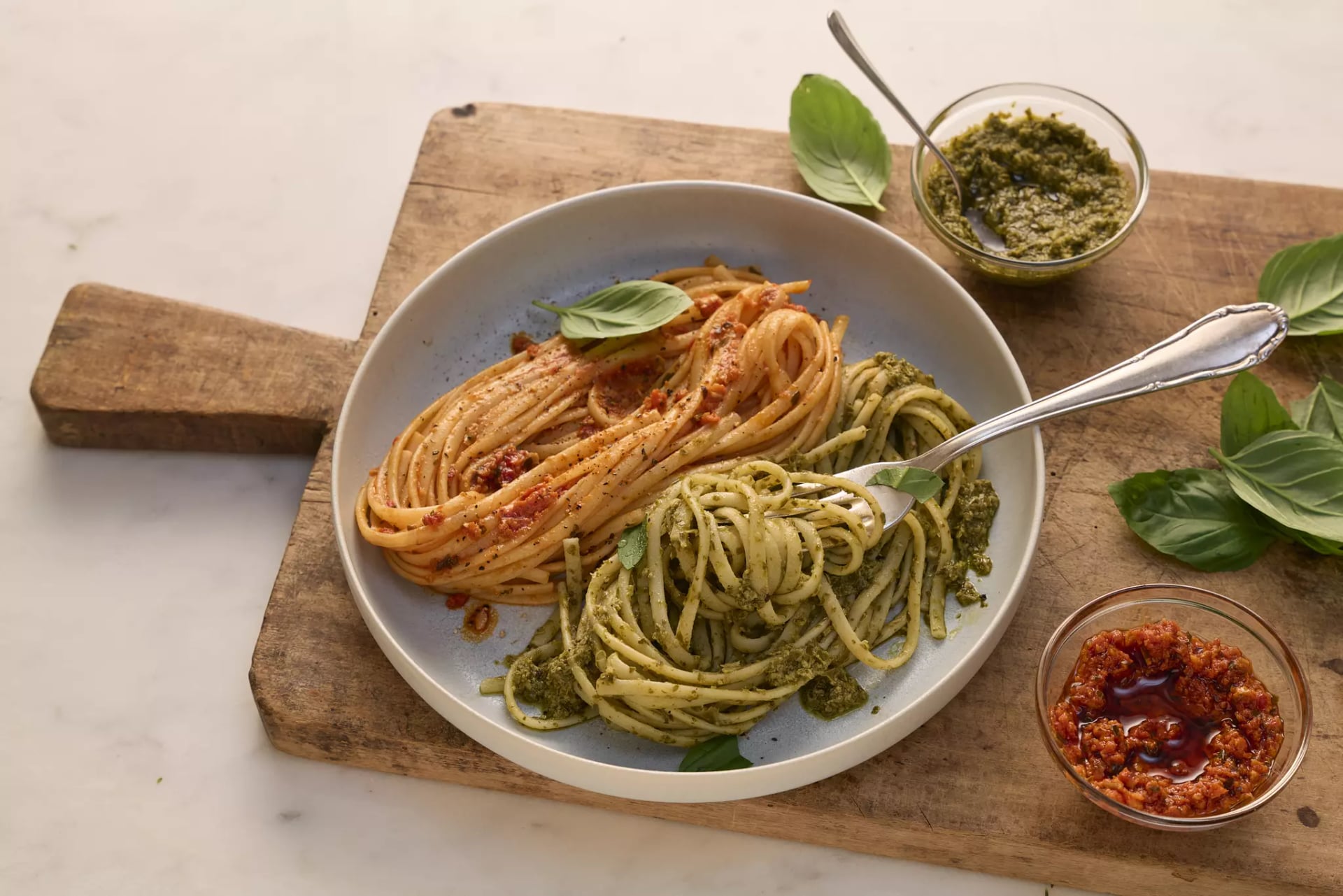 Ein flacher, weißer Teller mit zwei Portionen Spaghetti, die jeweils mit unterschiedlichen Pestos serviert werden. Links liegen die roten Spaghetti Pesto mit roten, leicht stückigen Tomaten, mit schwarzen Pfefferkörnern und Kräutern garniert. Rechts sind die grünen Spaghetti mit frischen Basilikumblättern garniert. Neben dem Teller befinden sich zwei kleine Schälchen: eines mit dem grünen Pesto und eines mit der roten Pesto. Eine silberne Gabel liegt auf dem Teller. Der Teller steht auf einem rustikalen, hellbraunen Holztisch, auf dem auch einige frische Basilikumblätter verteilt sind.