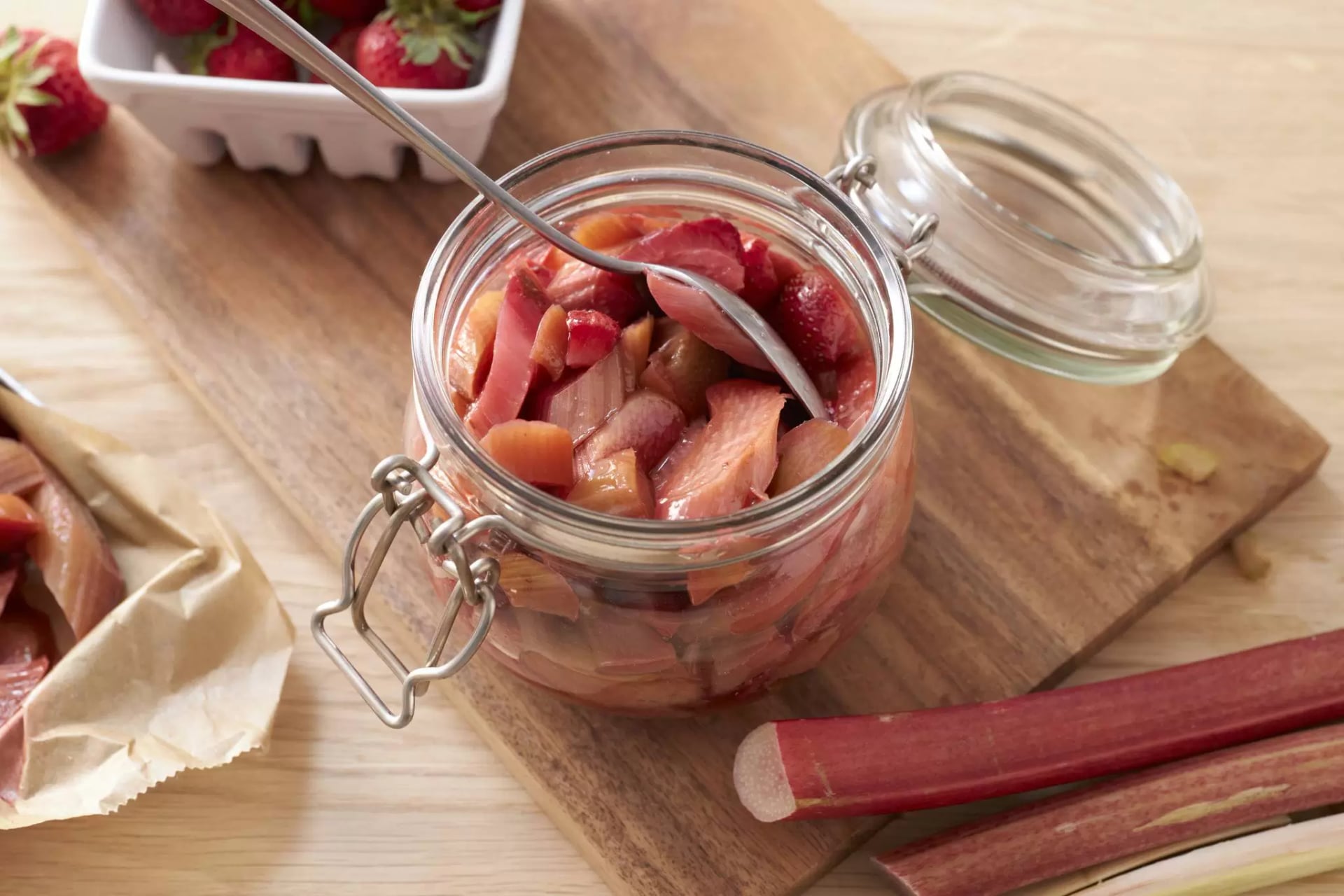 Offenes Glas mit geschnittenem Rhabarber und einem Löffel auf einem Holzbrett, daneben frische Erdbeeren in einer weißen Schale und zwei Rhabarberstangen