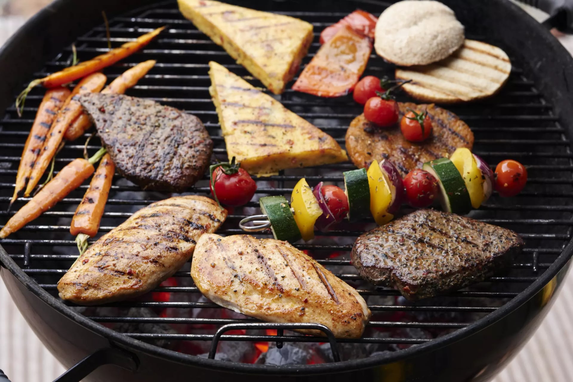 Auf dem Gitter eines Kugelgrills liegen verschiedene gegrillte Gemüse, Käse- und Fleischvarianten.