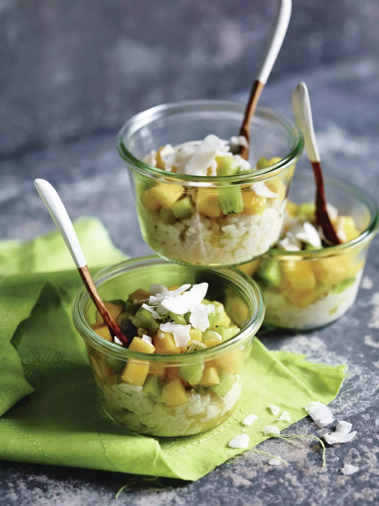 Drei Gläser mit Milchreis, Mango- und Kiwistücken, garniert mit Kokosraspeln und jeweils einem Löffel.