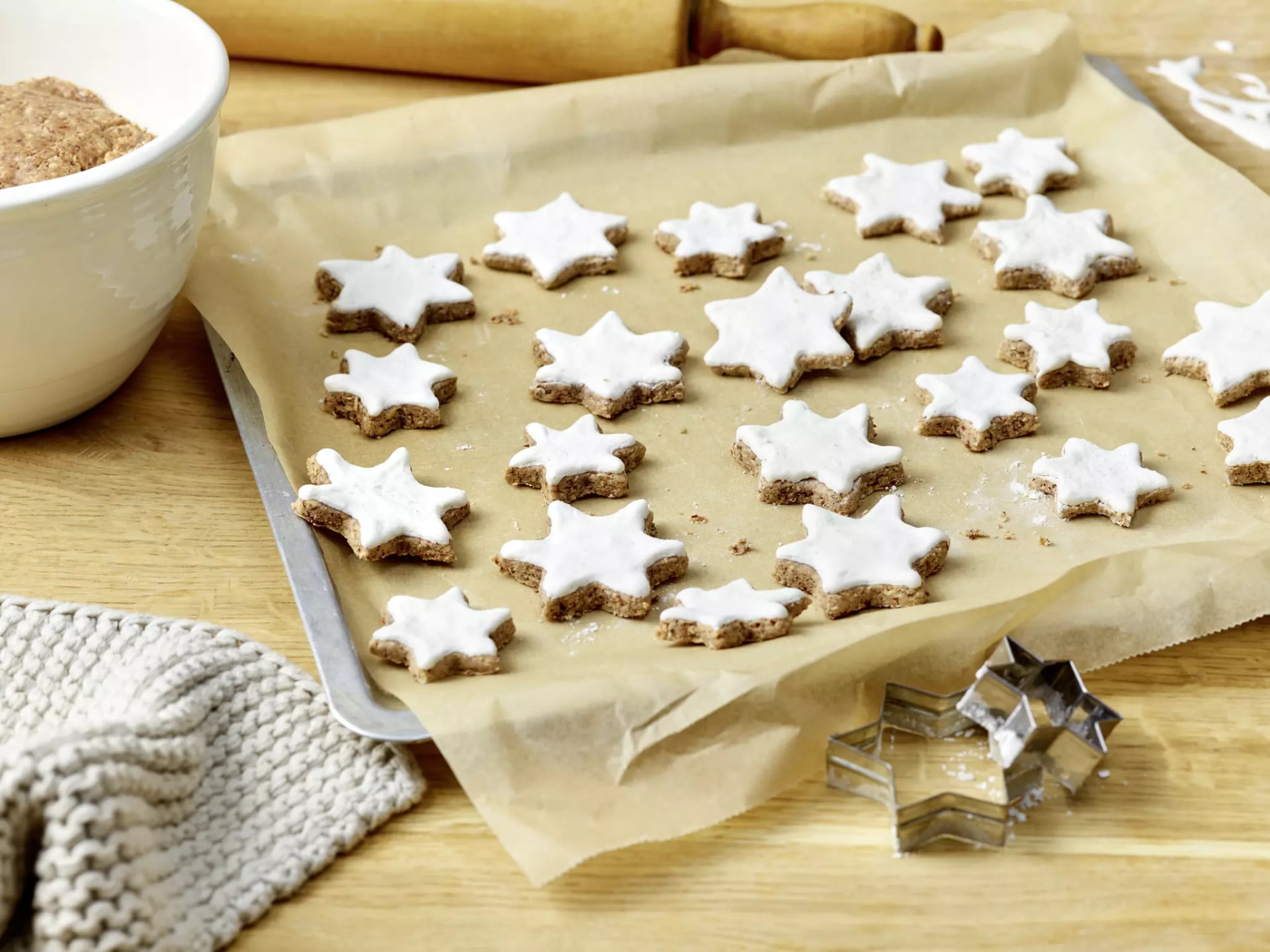 Backblech mit sternförmigen, mit Zuckerguss bedeckten Plätzchen auf Backpapier und zwei Metall-Ausstechformen in Sternform auf Holzuntergrund