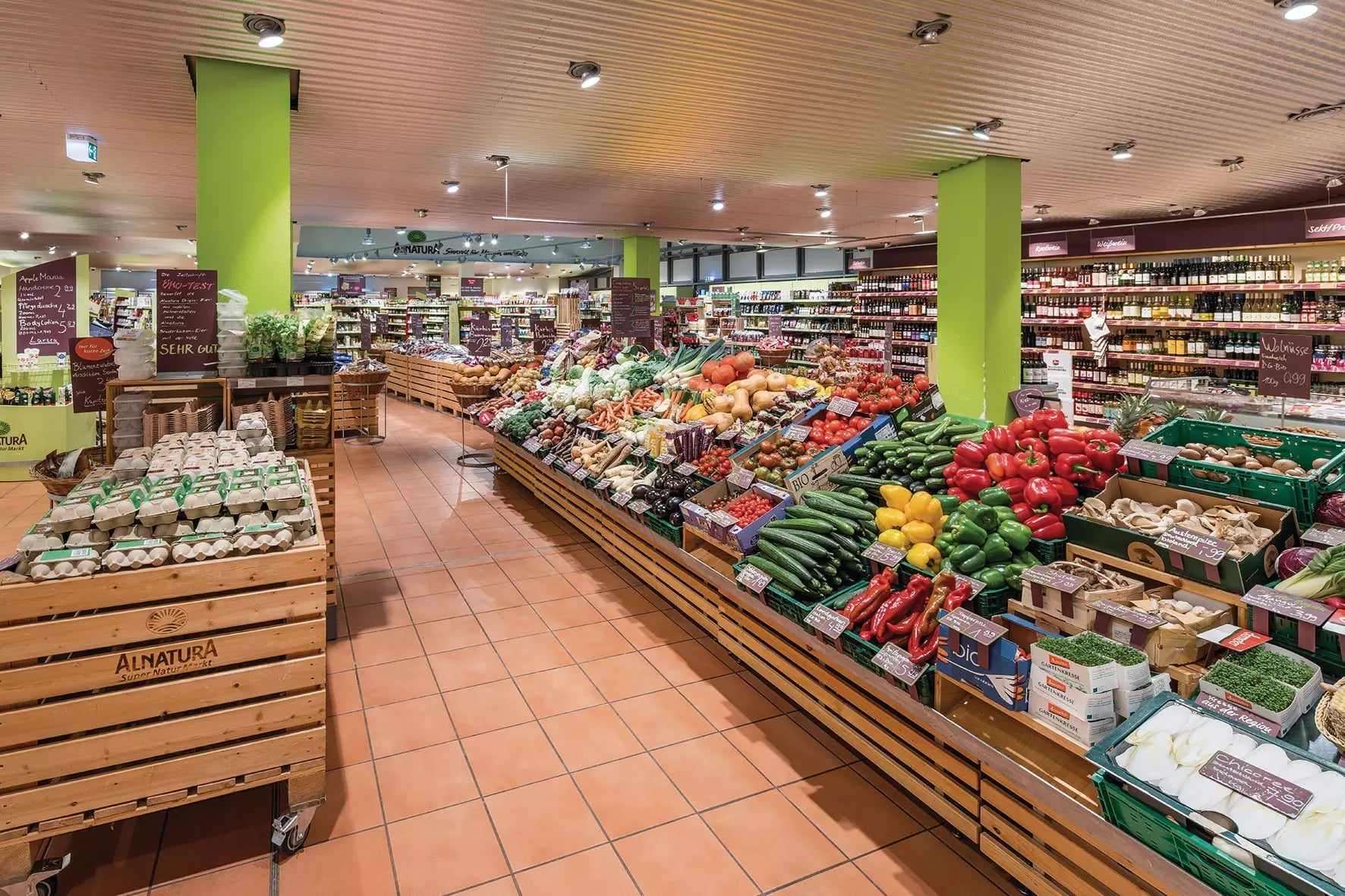 Verkaufsraum des Alnatura Super Natur Markts in Berlin mit Obst- und Gemüsestand, Holzkisten mit Eiern und grünen Säulen.