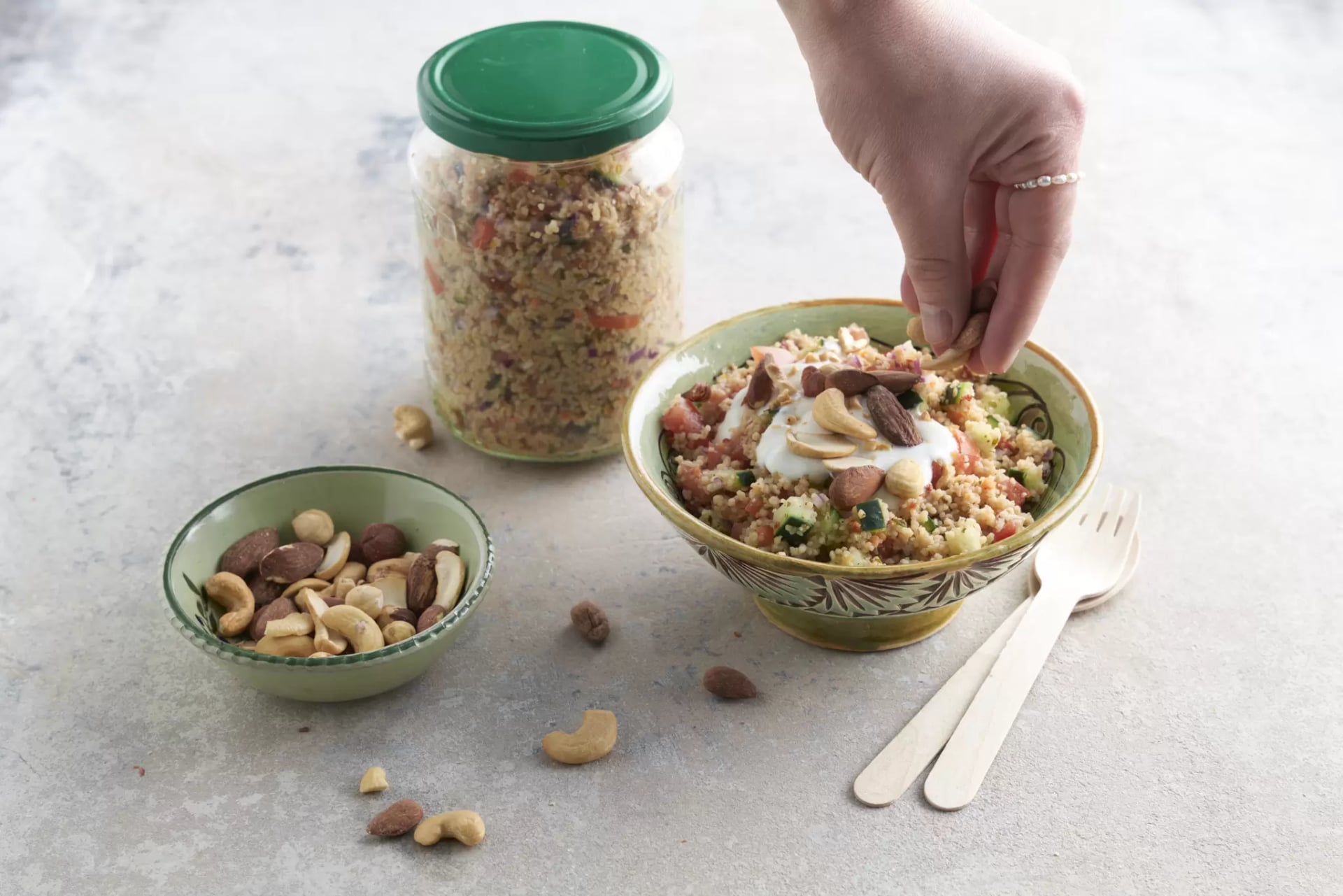 Schüssel mit Taboulé-Salat, Joghurt und Nüssen, daneben kleine Schale mit Nüssen und ein Glas mit grünem Deckel im Hintergrund, Hand streut Nüsse auf den Salat, helle Oberfläche mit zwei Holzbesteckteilen.