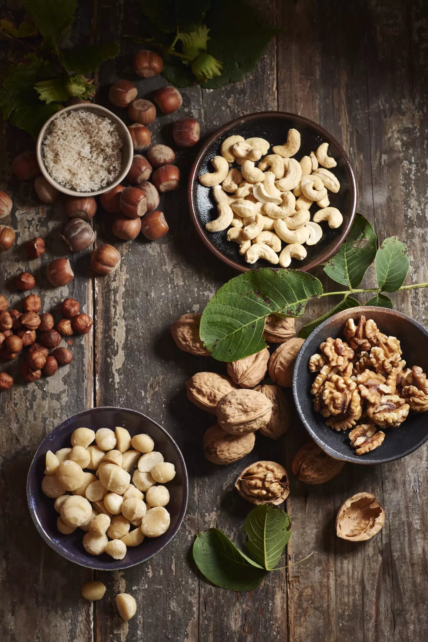 Verschiedene Nüsse in Schalen und mit Schalen auf rustikalem Holztisch, darunter Cashewkerne, Walnüsse, Haselnüsse und Macadamianüsse, umgeben von zwei grünen Blättern.