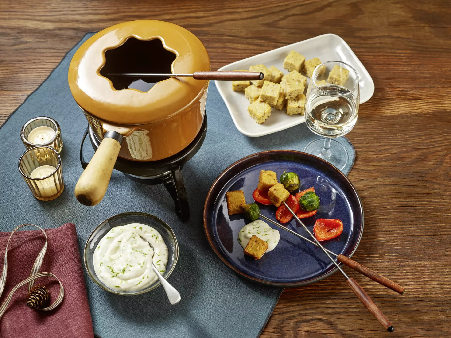 Fondue-Set mit orangem Topf, Holzgriff, Teller mit Brotstücken, Schale mit Dip, Glas Weißwein und zwei Fonduegabeln auf Holztisch