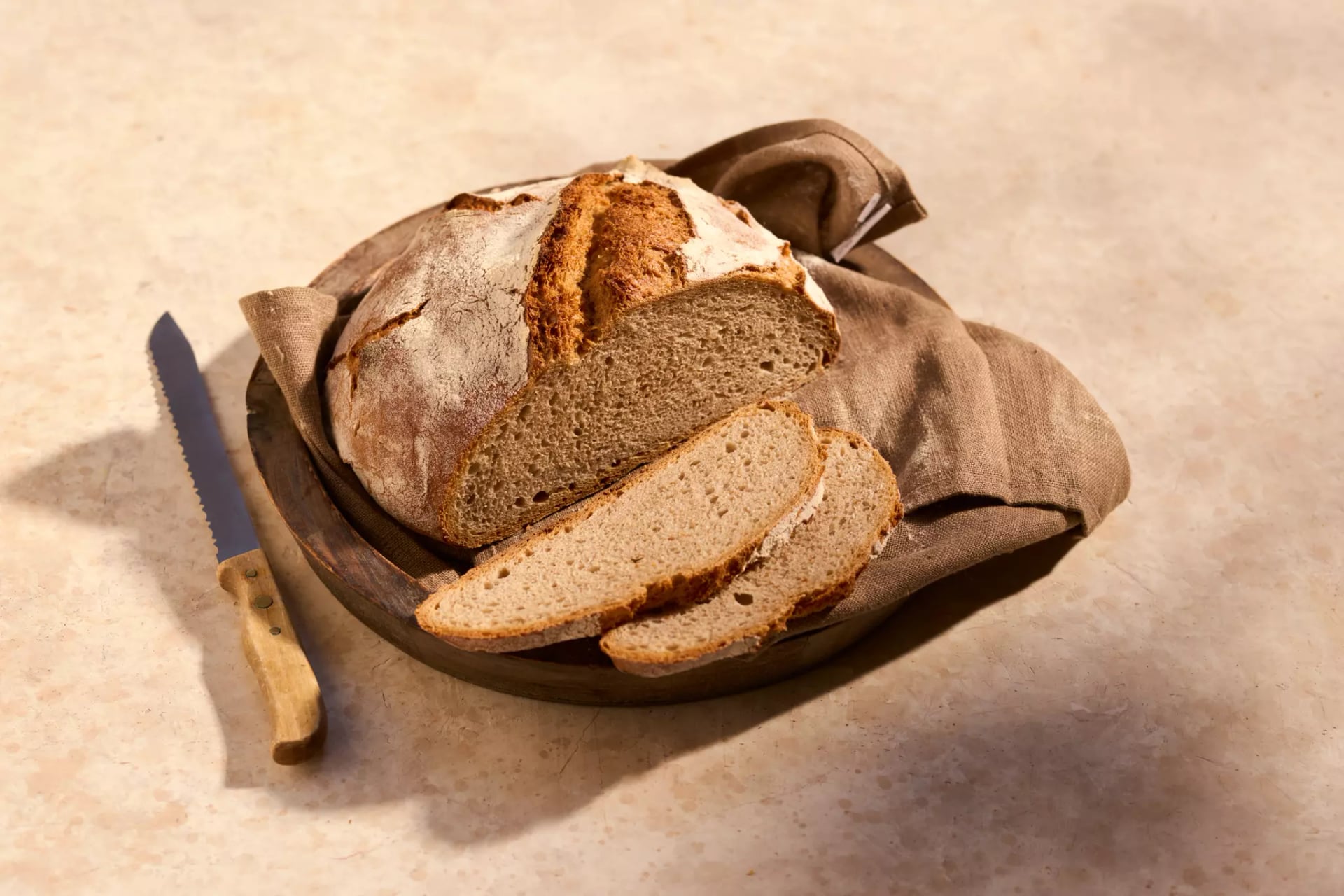 Angeschnittenes No-Knead_Brot mit zwei Brotscheiben in einer Holzschale, daneben liegt ein Brotmesser.