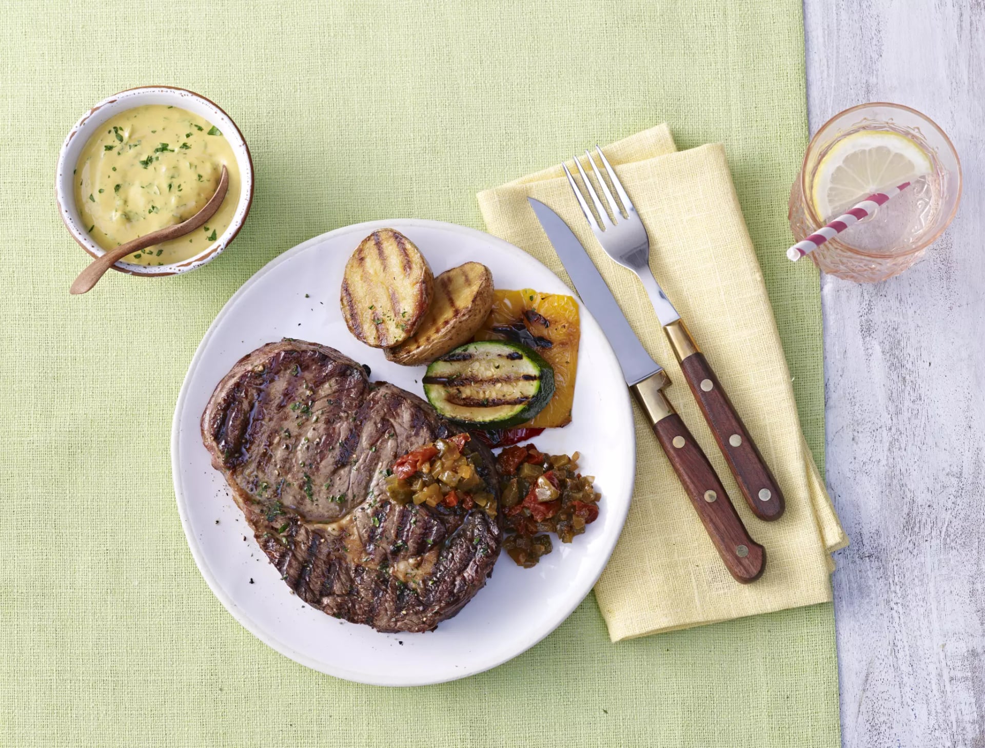Gegrilltes Steak mit gegrilltem Gemüse auf weißem Teller neben Besteck auf gelbem Stoffserviette und Glas mit Zitronenscheibe und Strohhalm.