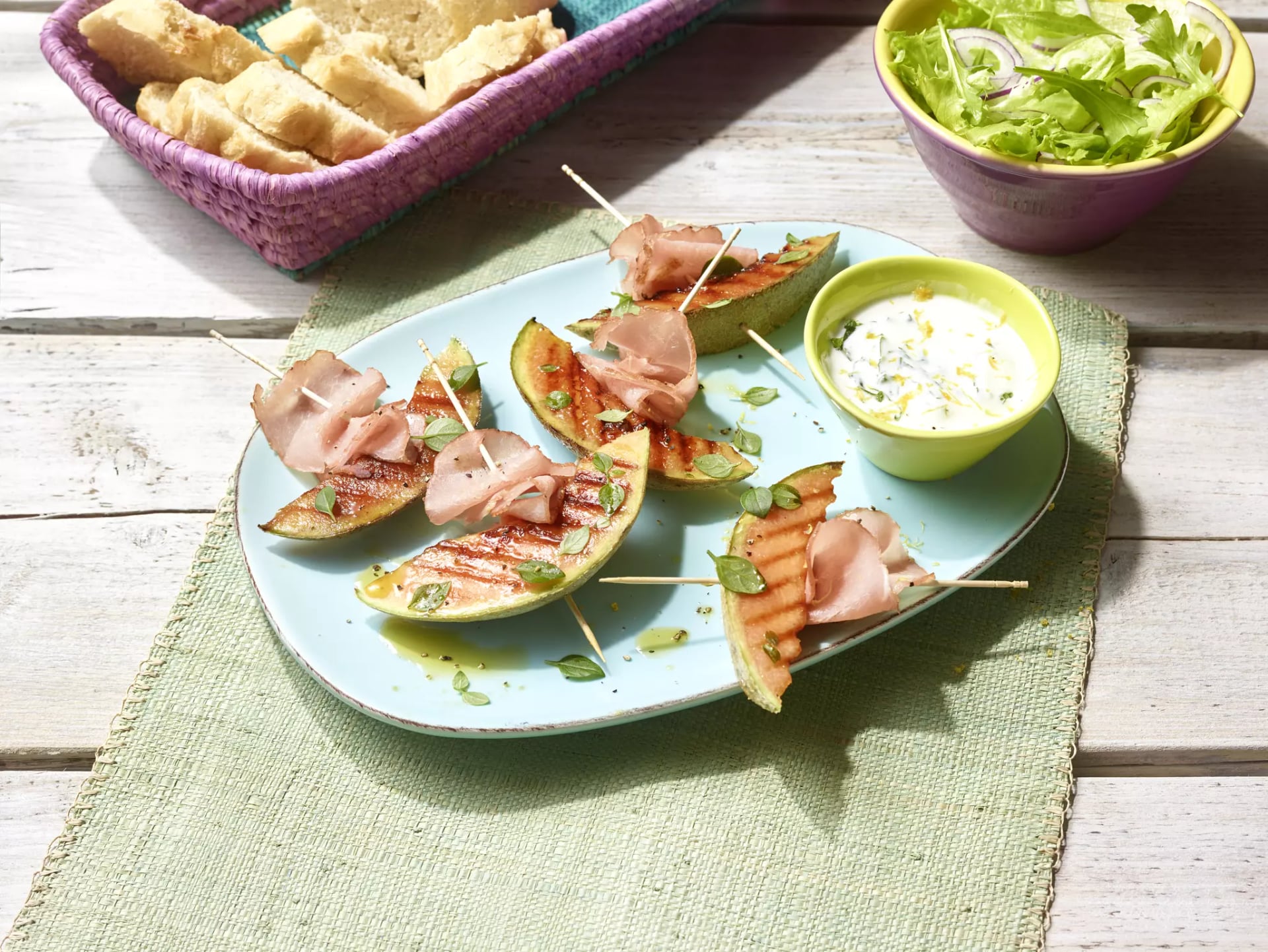 Gegrillte Melonenspalten mit Schinken auf einem Teller mit Dip und Salat im Hintergrund.