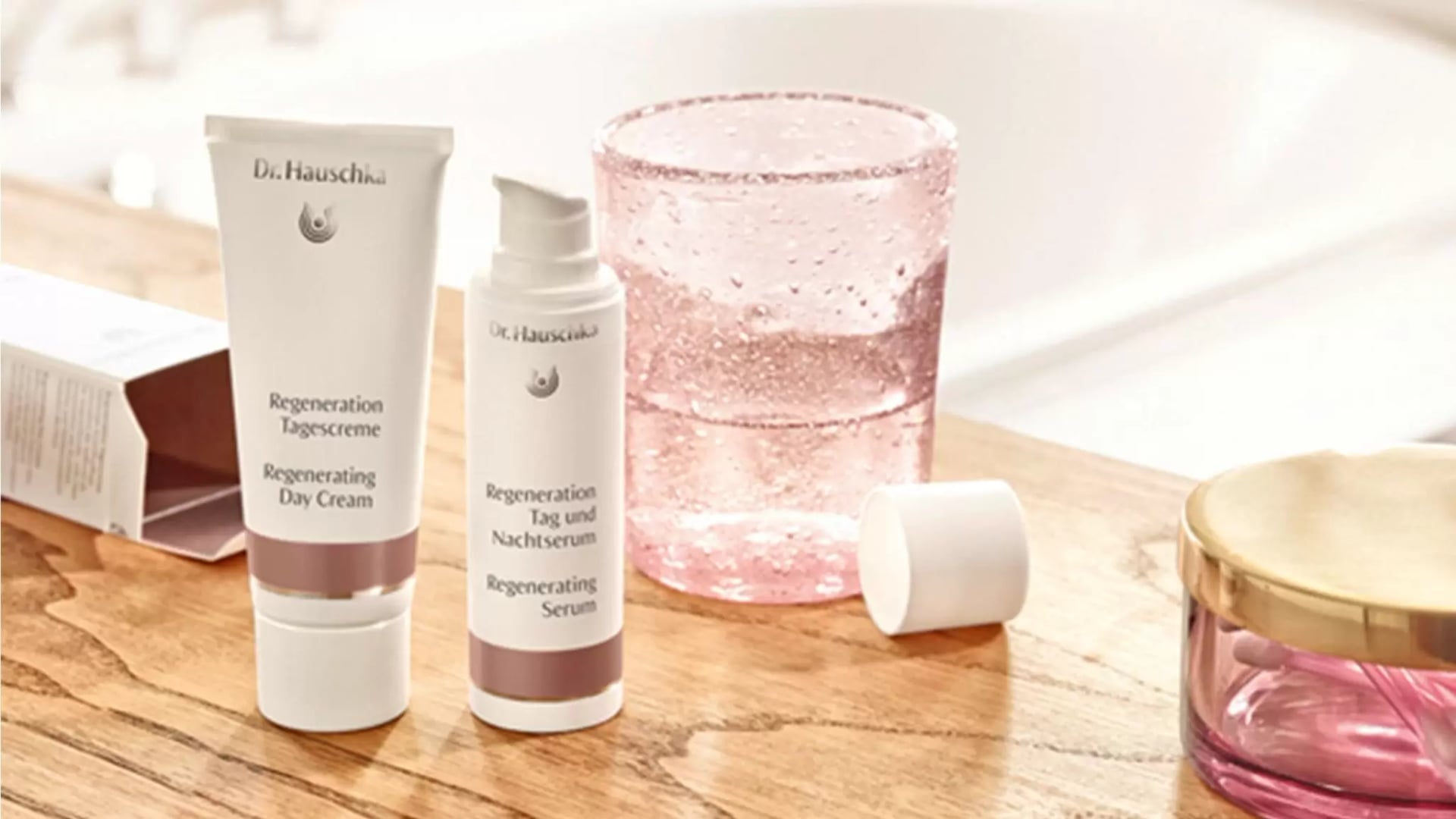 Zwei Dr.-Hauschka-Pflegeprodukte, eine Regenerations-Tagescreme und ein Regenerations-Tag-und-Nachtserum, stehen auf einem Holztisch neben einem rosafarbenen Glas und einem kleinen runden Behälter mit goldenem Deckel.