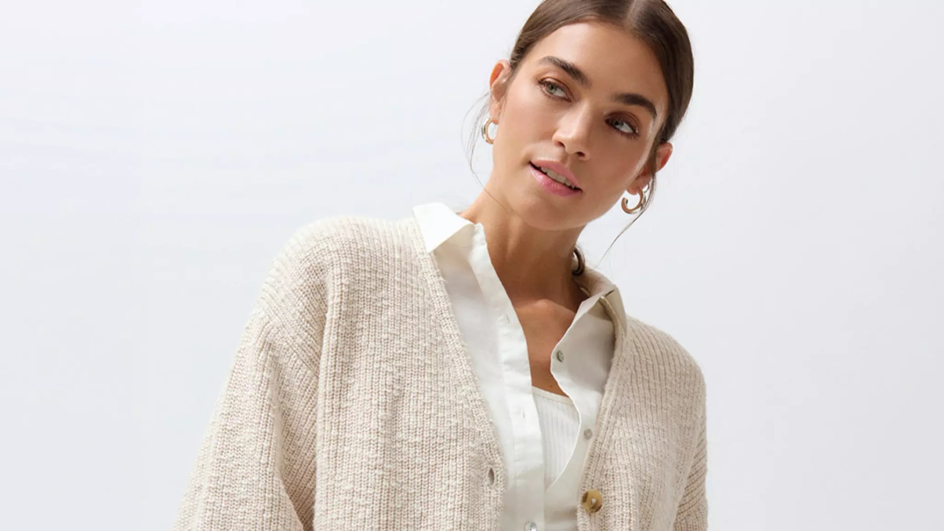 Frau mit zurückgebundenem Haar trägt weiße Bluse und grob gestrickte beige Strickjacke mit Knöpfen.