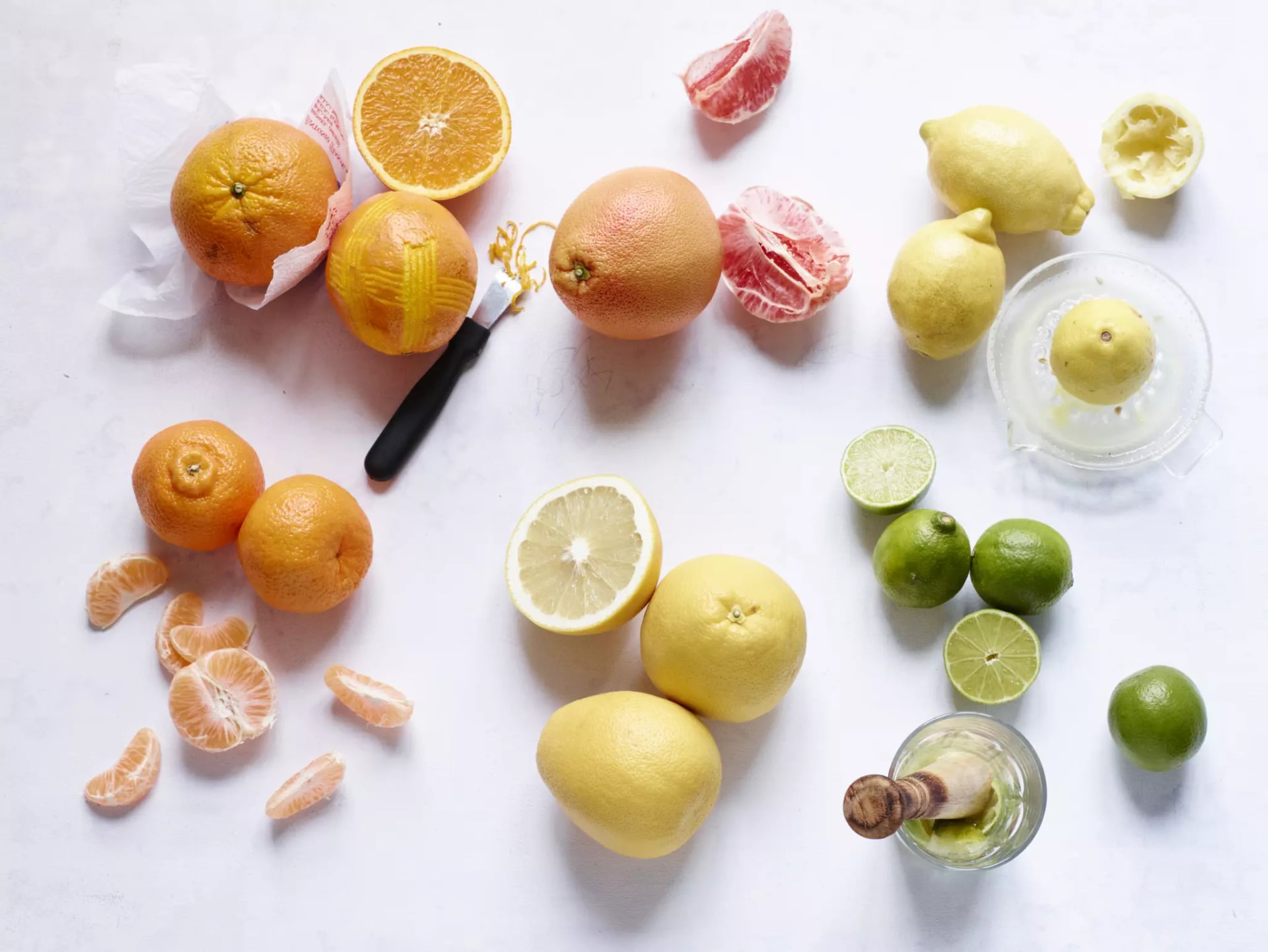 Verschiedene Zitrusfrüchte, darunter Orangen, Grapefruits, Zitronen und Limetten, teils ganz, teils geschält oder halbiert, mit einem Entsafter und einem Messer auf weißem Untergrund.
