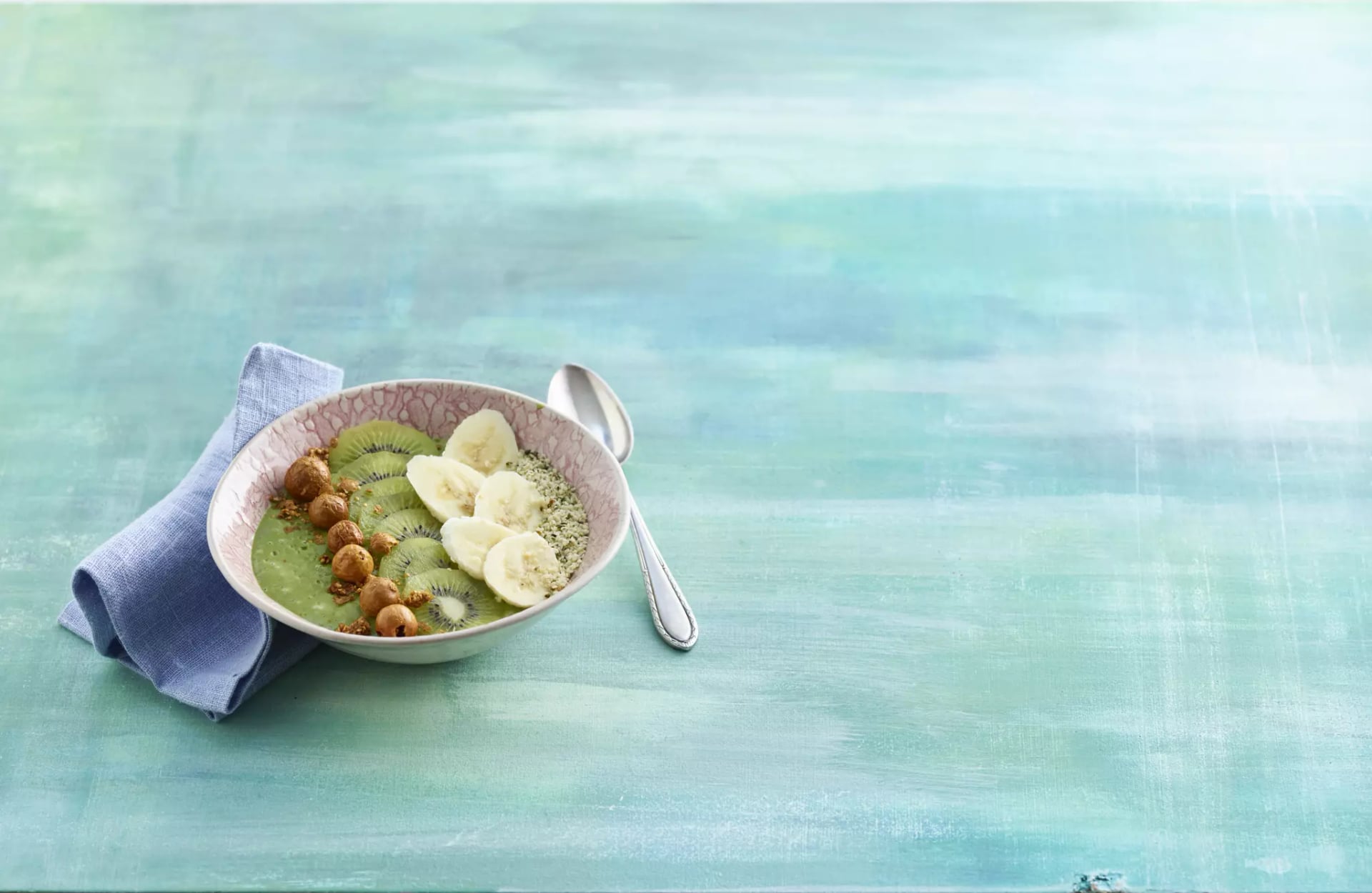 Schale mit grüner Smoothie-Basis, garniert mit Bananenscheiben, Kiwischeiben, Haselnüssen und Samen, daneben ein Löffel und ein blaues Tuch auf türkisfarbenem Untergrund.