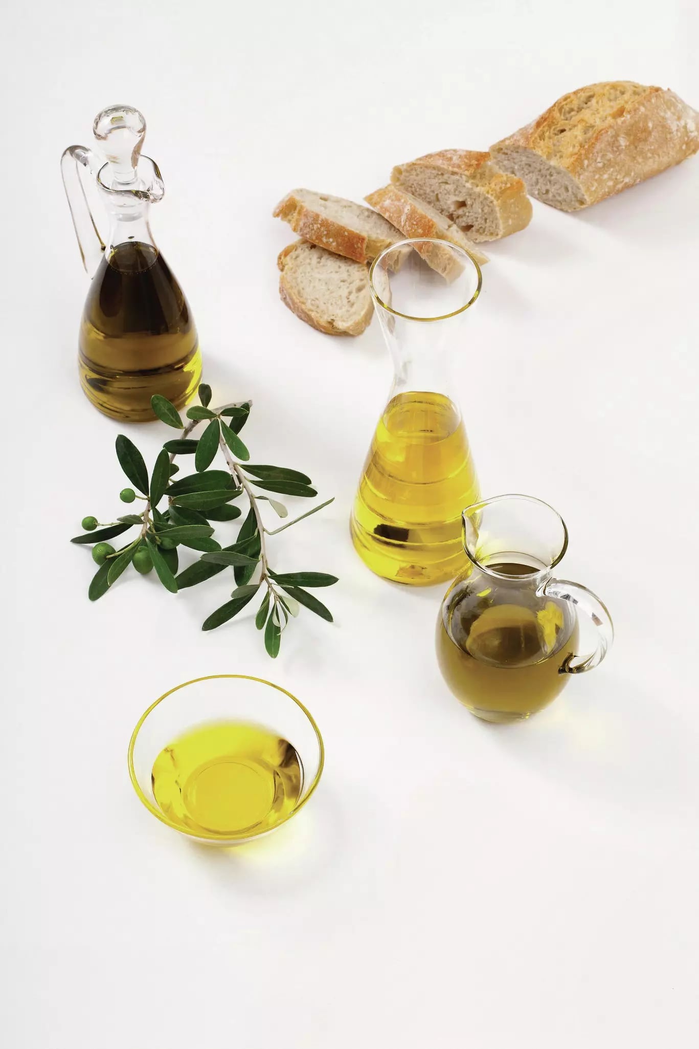 Zwei Öl in Glasflaschen und ein helles Öl in einer Glasschale. Daneben liegt ein aufgeschnittenes Baguette und ein Oliven-Zweig.