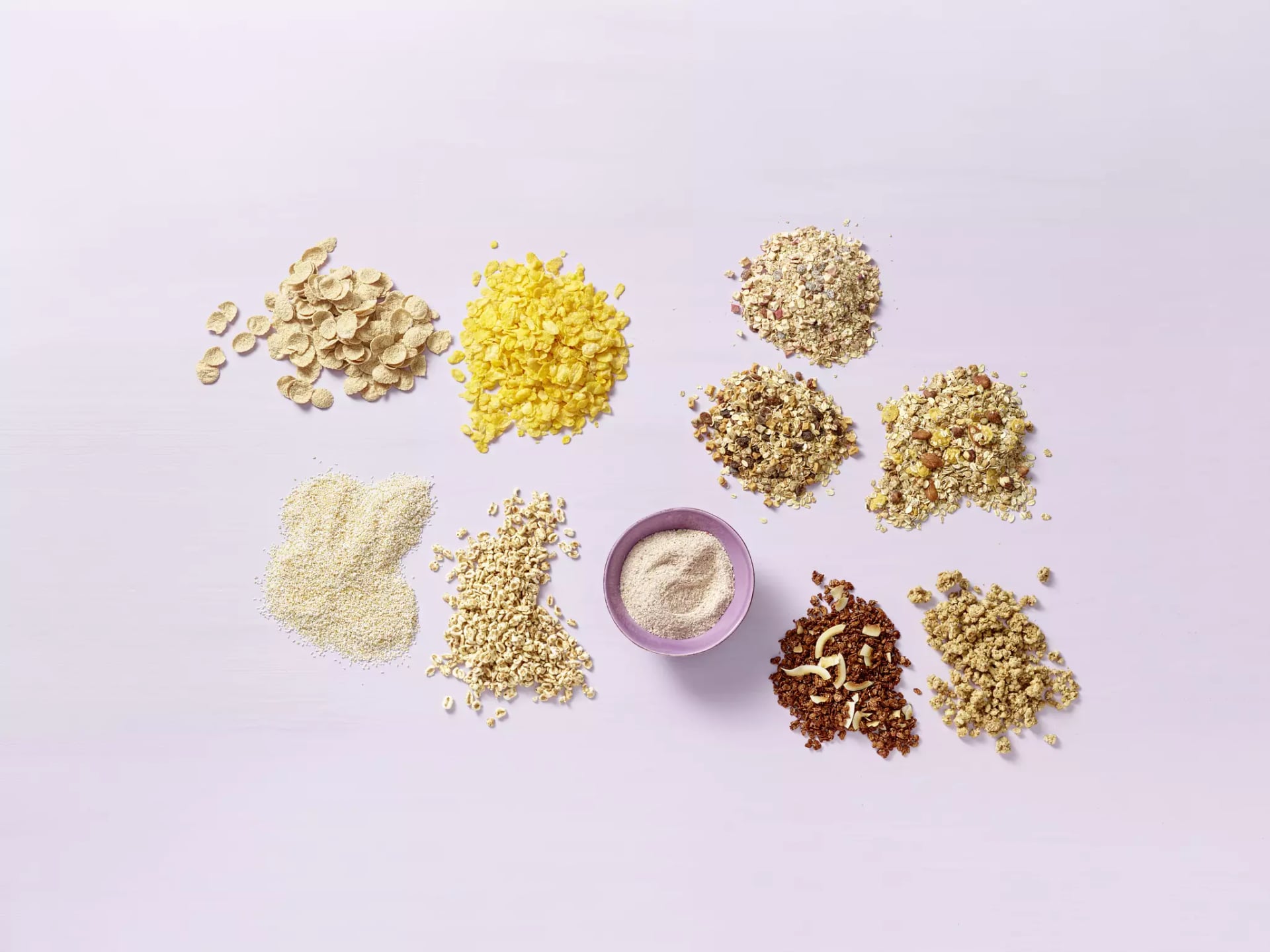 Mehrere kleine Haufen verschiedener Getreideflocken und ein lila Becher mit Amaranth auf hellem Untergrund.