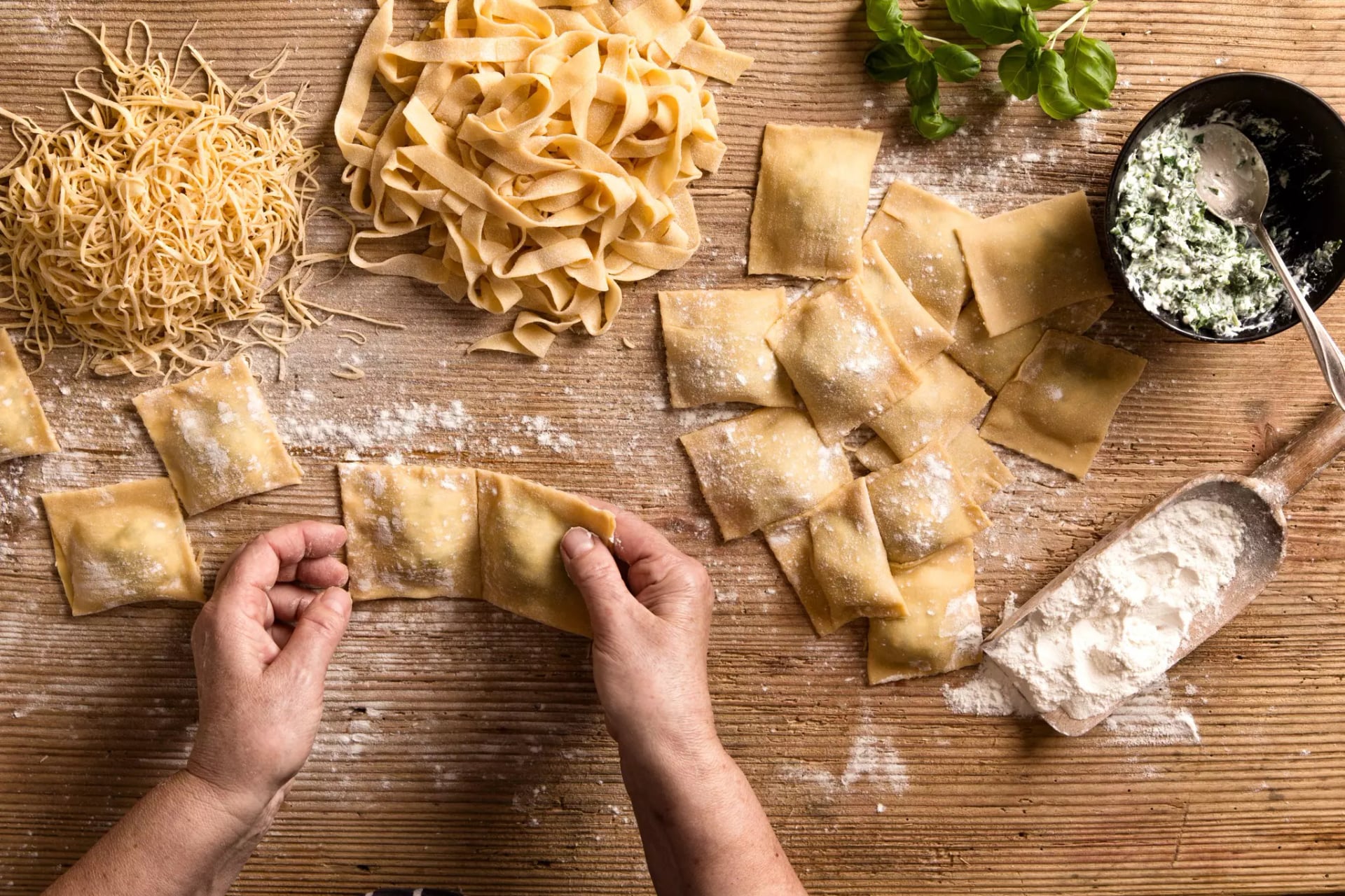 Zwei Hände formen Ravioli. Drum herum sieht man Mehl, Basilikum, fertige Ravioli sowie Tagiatelle und Spaghetti aus frischem Teig sowie eine Füllung in einer schwarzen Schale.