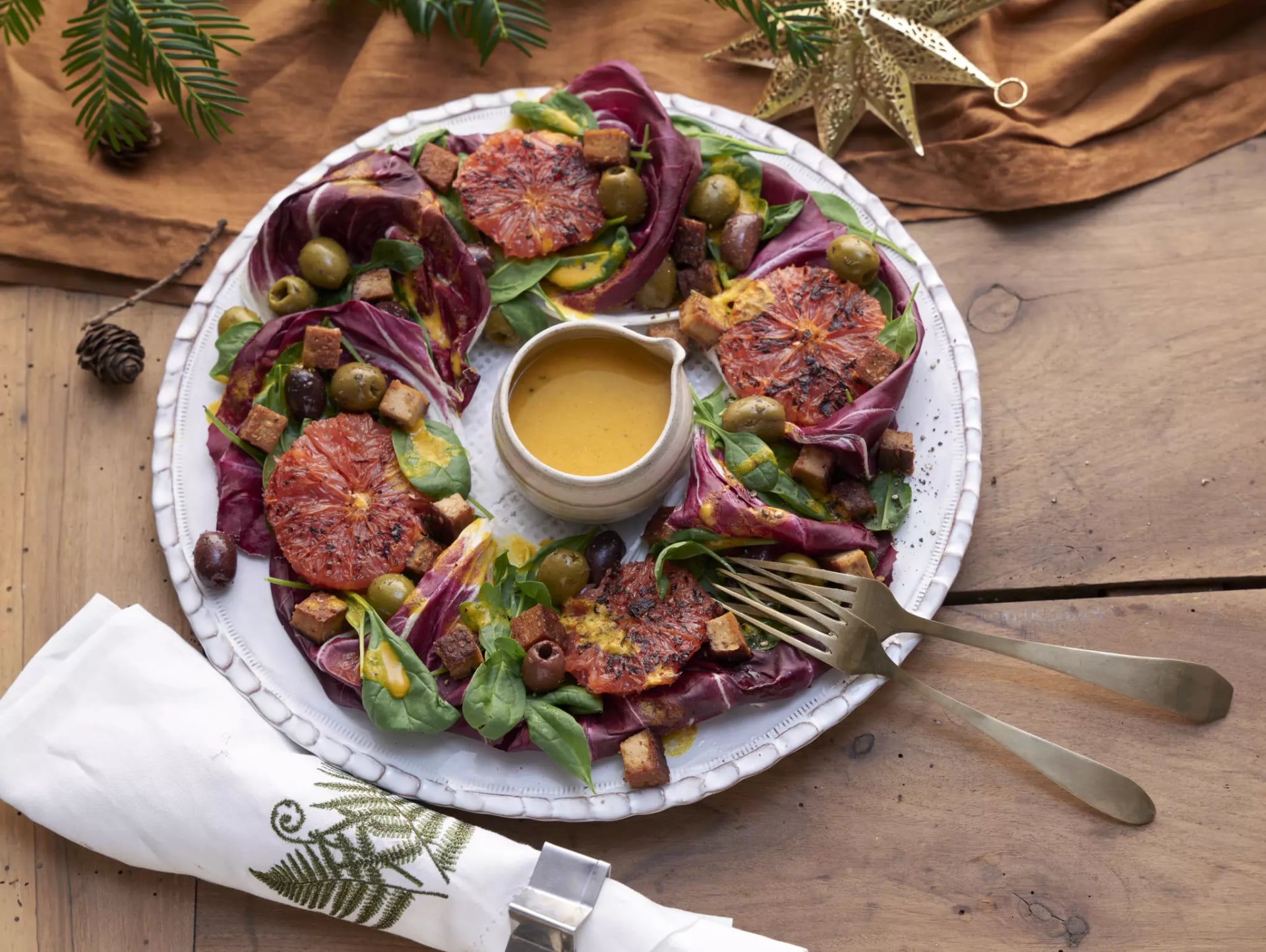 Ein dekorativ angerichteter Salatkranz auf einem weißen Keramikteller. Der Salat besteht aus violetten Radicchio-Blättern, frischem Babyspinat, gebratener Grapefruit und knusprigen Tofuwürfeln. In der Mitte des Kranzes steht ein kleines Kännchen mit einem gelben Dressing. Das Ganze ist auf einem Holztisch platziert, dekoriert mit einer Stoffserviette, goldenem Besteck, Tannenzweigen und einem goldenen Weihnachtsstern.