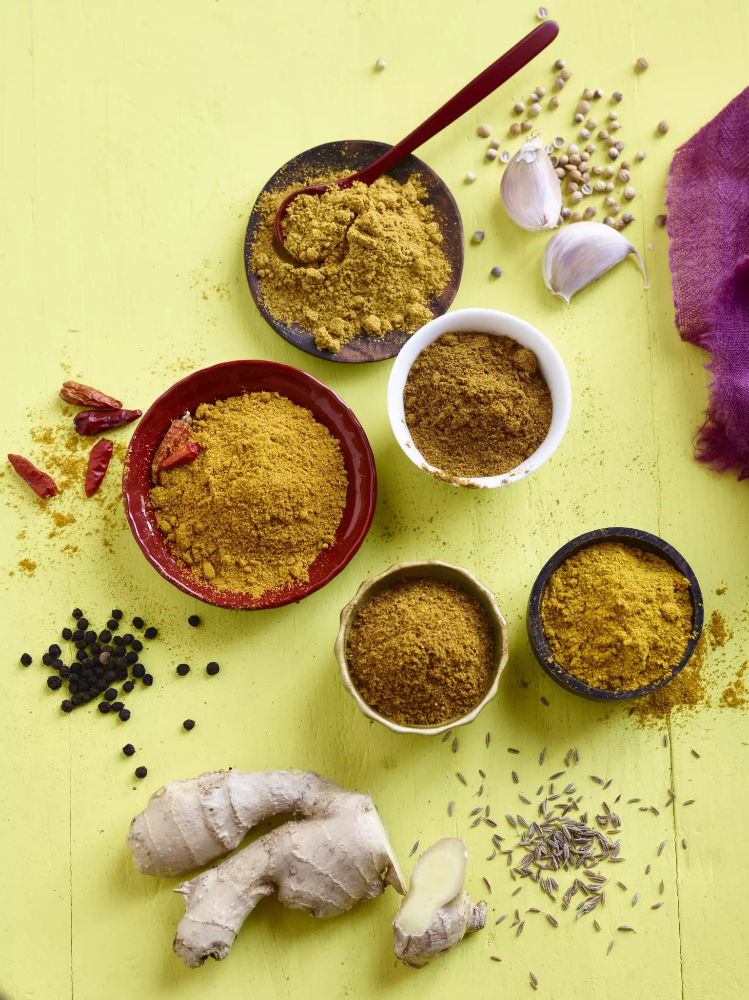 Verschiedene Currygewürze in kleinen Schälchen auf einem geblichen Untergrund. Daneben liegen eine Ingwerknolle, Pfefferkörner und Knoblauch.