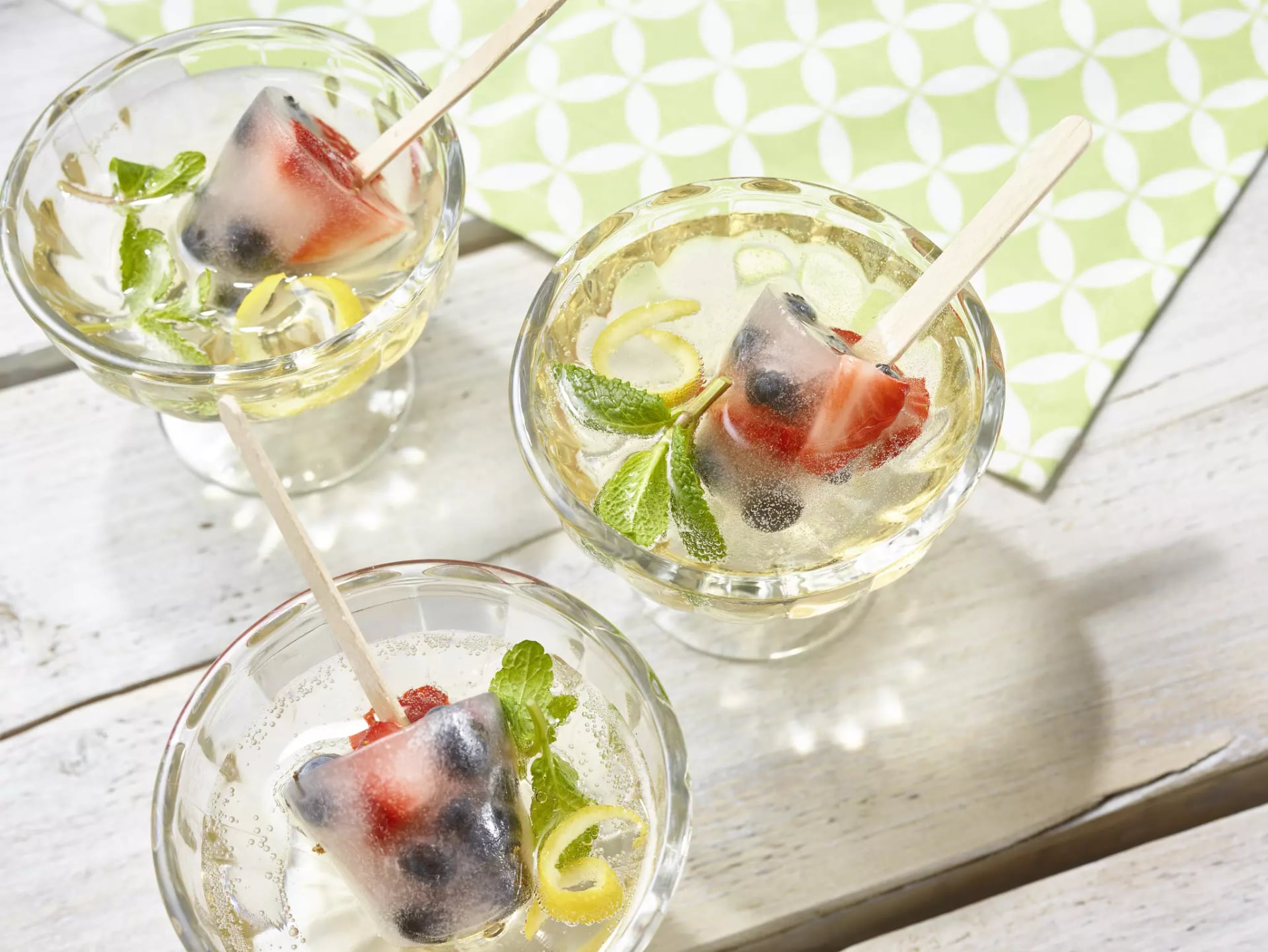 Drei Gläser mit Getränk, Eiswürfeln mit Beeren und Minzblättern, auf hellem Holztisch mit grün-weiß gemustertem Tuch.