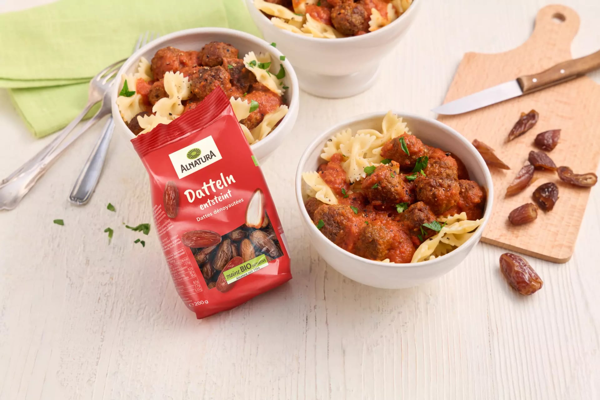Schale mit Farfalle-Nudeln und Fleischbällchen in Tomatensauce neben Packung Alnatura Datteln und Schneidebrett mit entsteinten Datteln