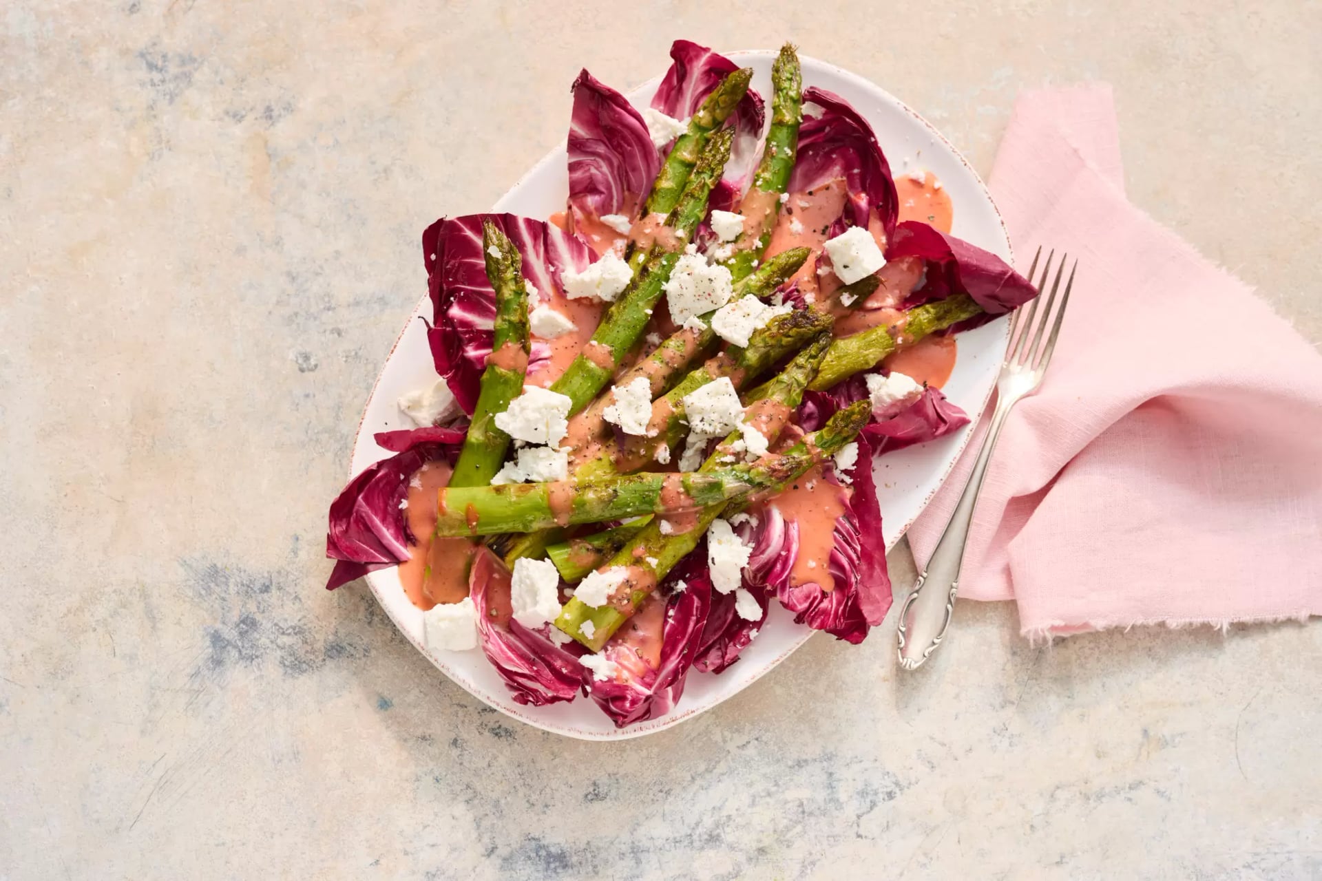 Grüner Spargel auf Radicchio-Blättern mit zerbröseltem Manouri-Käse auf weißem Teller neben silberner Gabel und rosa Stoffserviette.