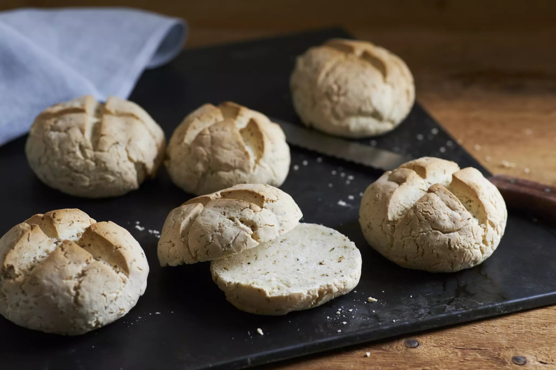 Sechs glutenfreie Kräuterbrötchen auf einem Brett. Eines der Brötchen ist aufgeschnitten.