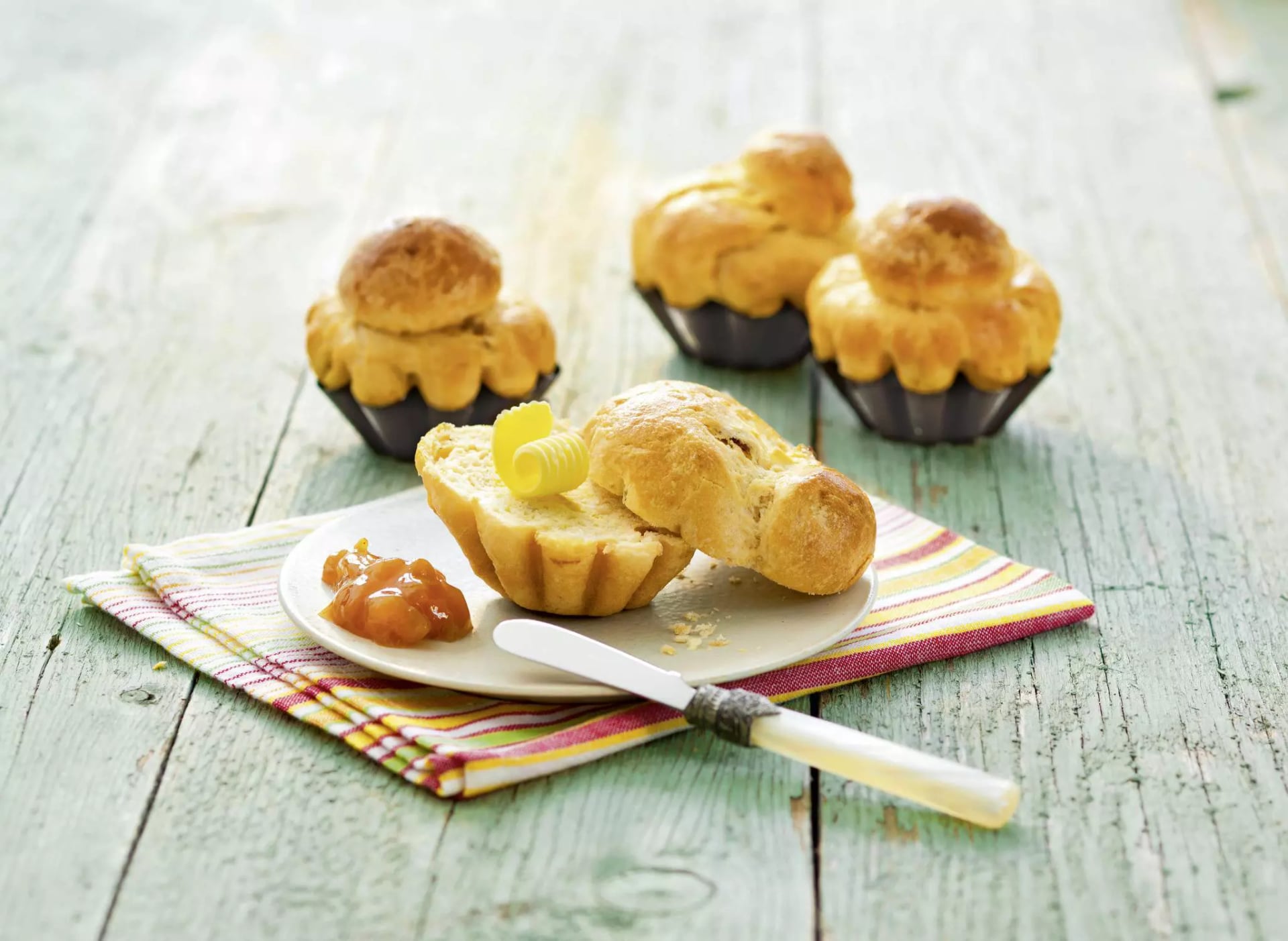 Aufgeschnittenes Brioche mit Butter bestrichen, daneben Marmelade. Im Hintergrund drei Brioches in den Backförmchen.