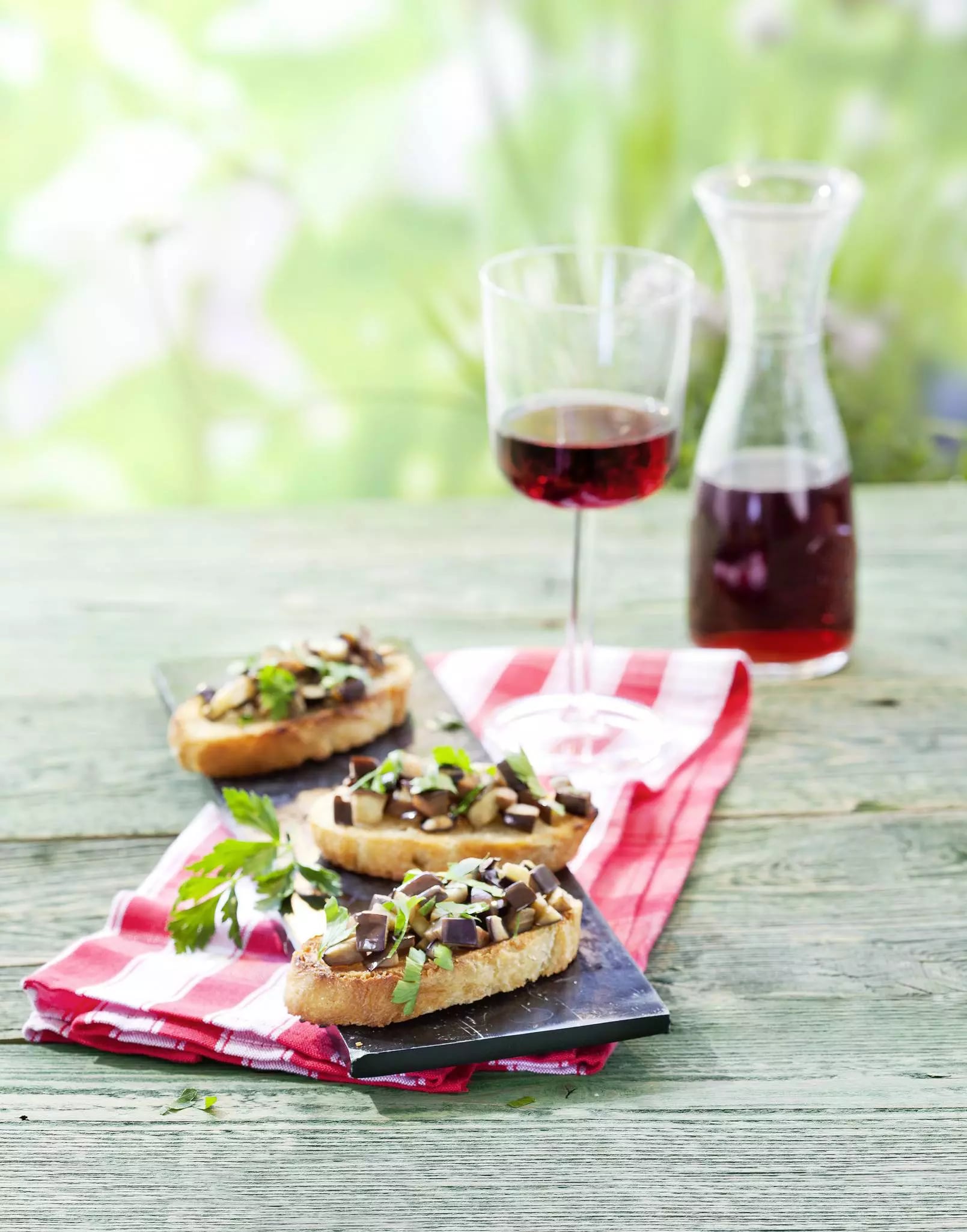 Drei mit Auberginen belegte Bruschetta auf schwarzer Platte mit rotem Weinglas und Karaffe auf rot-weiß kariertem Tuch vor grünem Hintergrund.