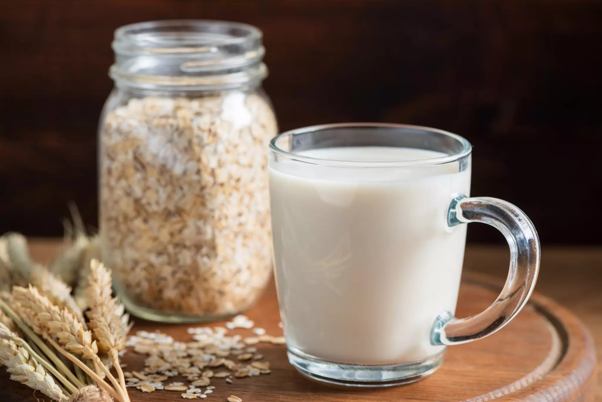 Eine Tasse in der sich Hafermilch befindet, daneben ein Glas Haferflocken und ein paar Gerstenhalme