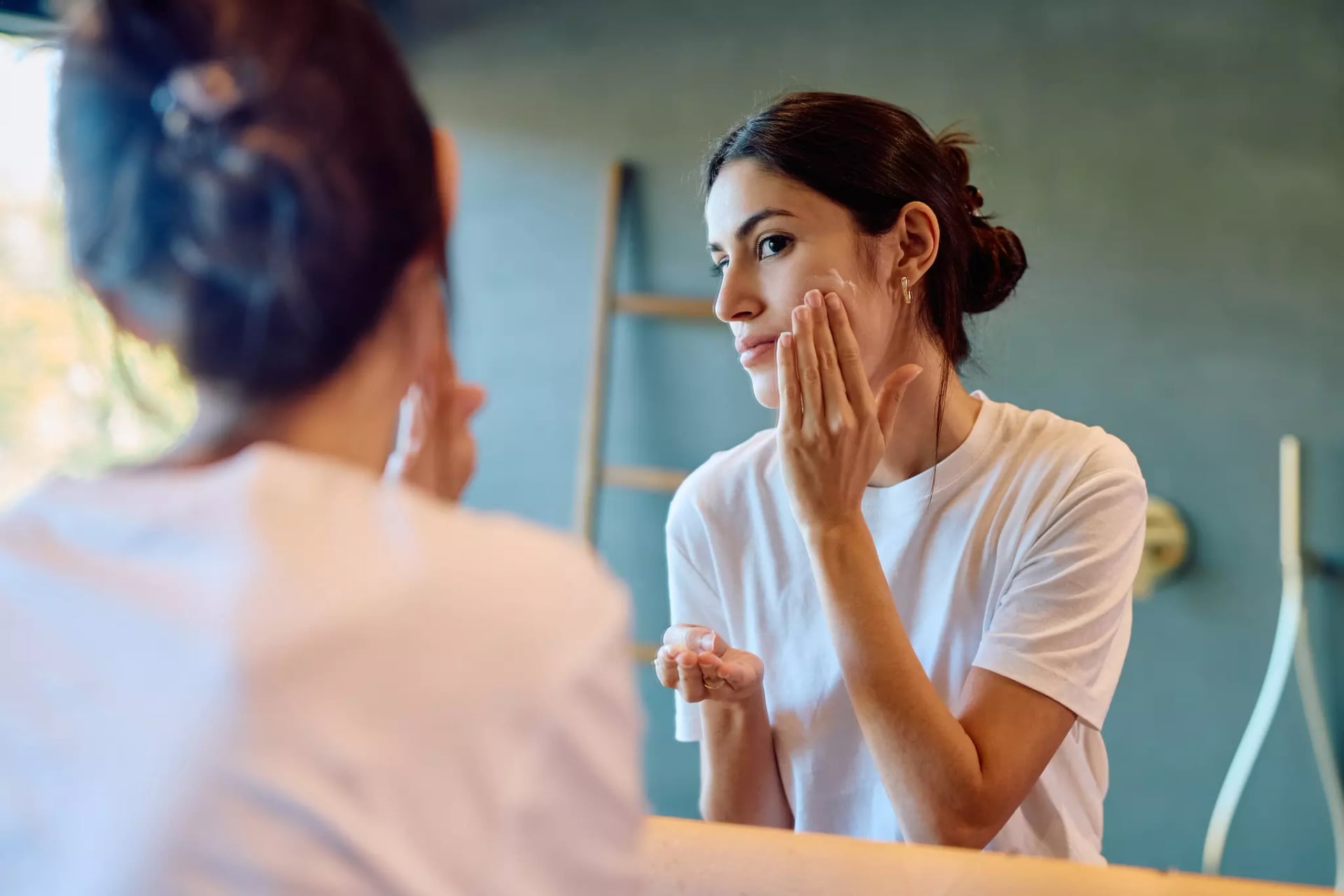 Eine brünette Frau steht vor dem Spiegel und reibt sich mit einer Hand Creme auf die Wange