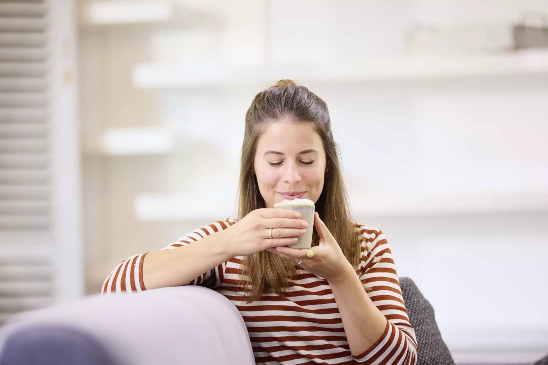 Frau mit langem Haar und gestreiftem Oberteil sitzt auf Sofa und hält eine Tasse mit beiden Händen nahe an ihr Gesicht