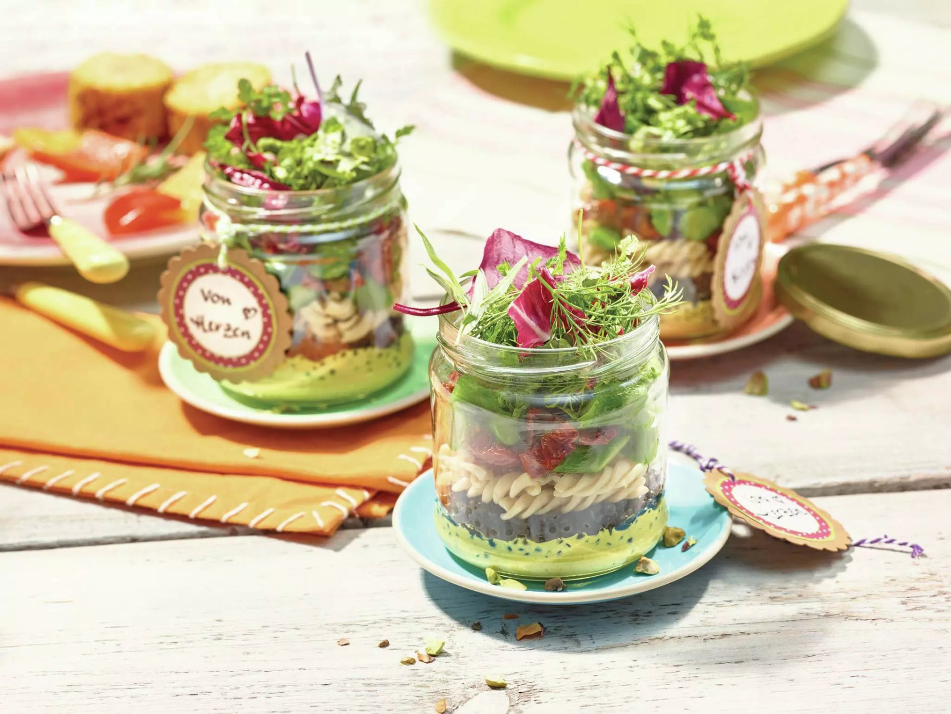 Drei Gläser voll mit cremigem Cury-Nudel-Linsen-Salat mit Salat, dekoriert mit kleinen Anhängern, auf Tellern und einem orangefarbenen Tuch auf einem Holztisch.