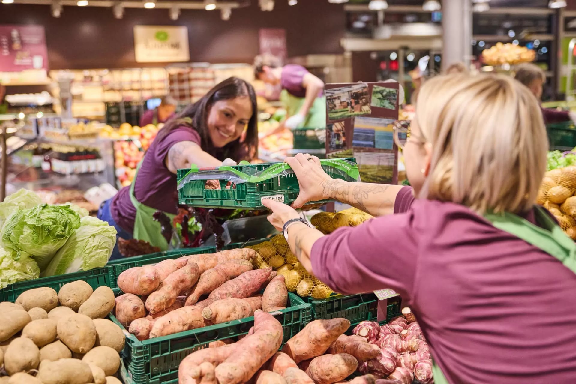 Mitarbeiterinnen im Alnatura Markt räumen die Obst und Gemüseabteilung ein und reichen sich eine Kiste über das Regal.