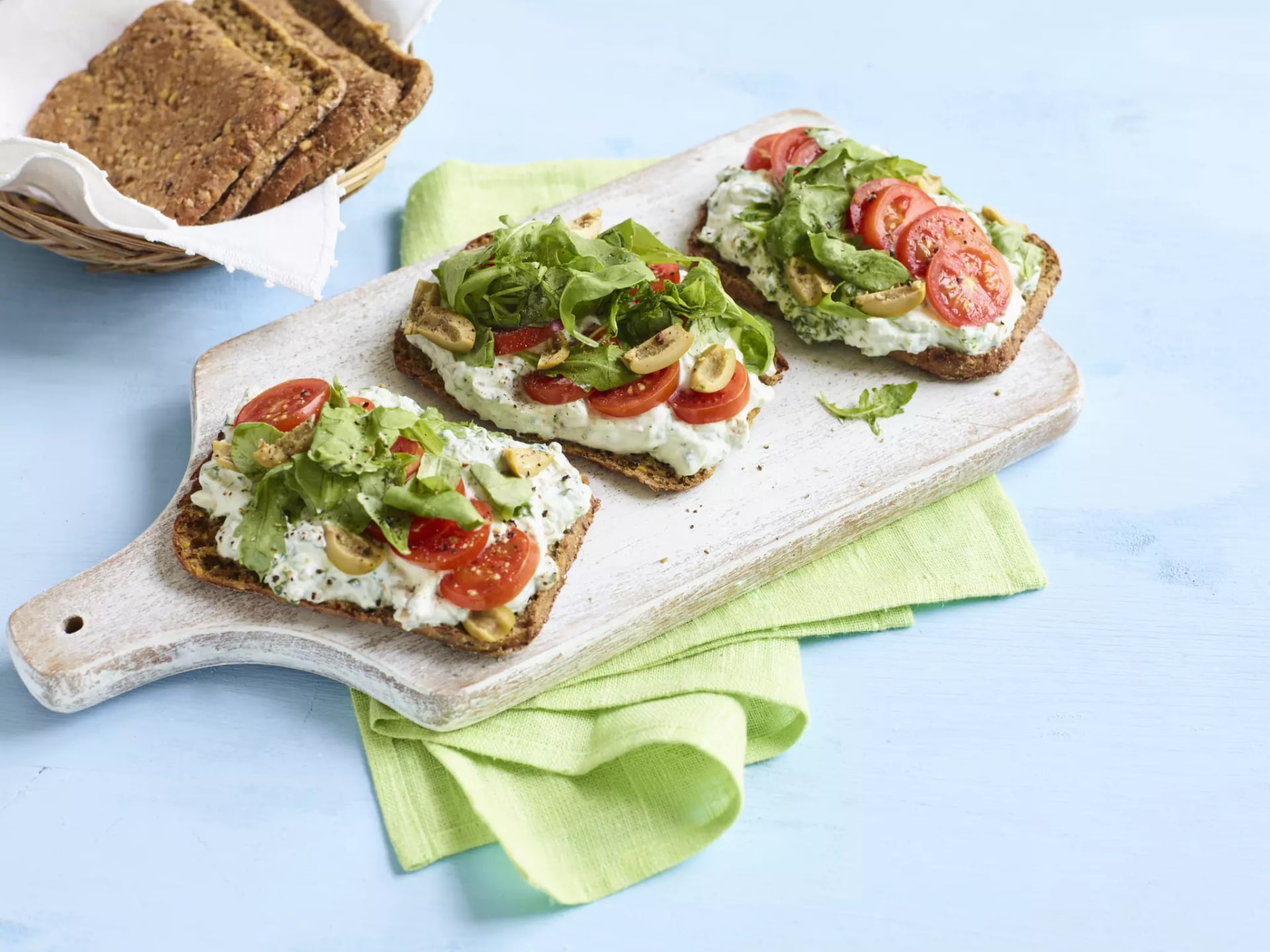 Drei belegte Vollkornbrotscheiben mit Frischkäse, Tomatenscheiben, Salat und Olivenscheiben auf einem weißen Holzbrett neben einem Brotkorb mit Vollkornbrot.