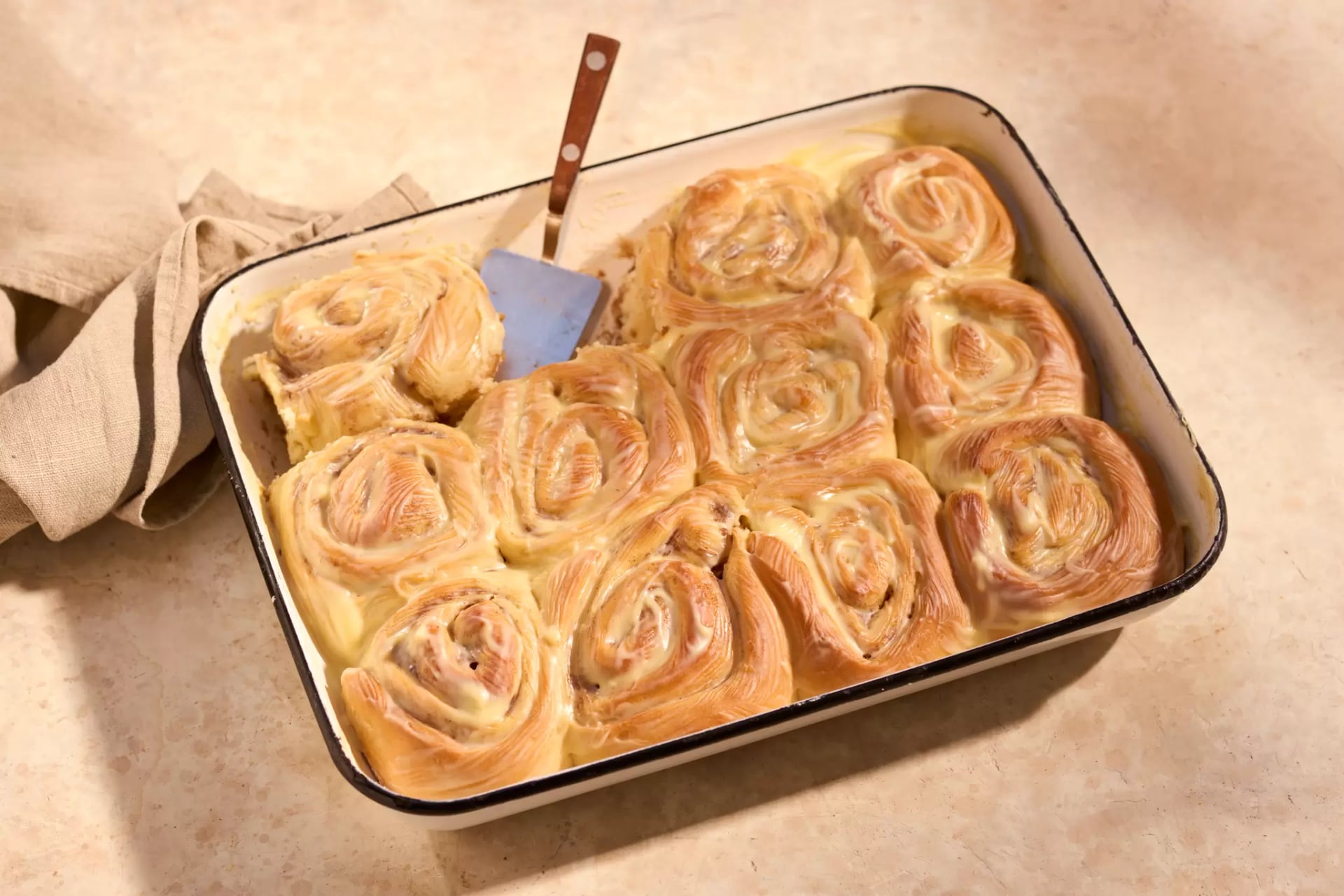 Mehrere Apfel-Zimtschnecken in einer rechteckigen Backform, daneben liegt ein helles Küchentuch.