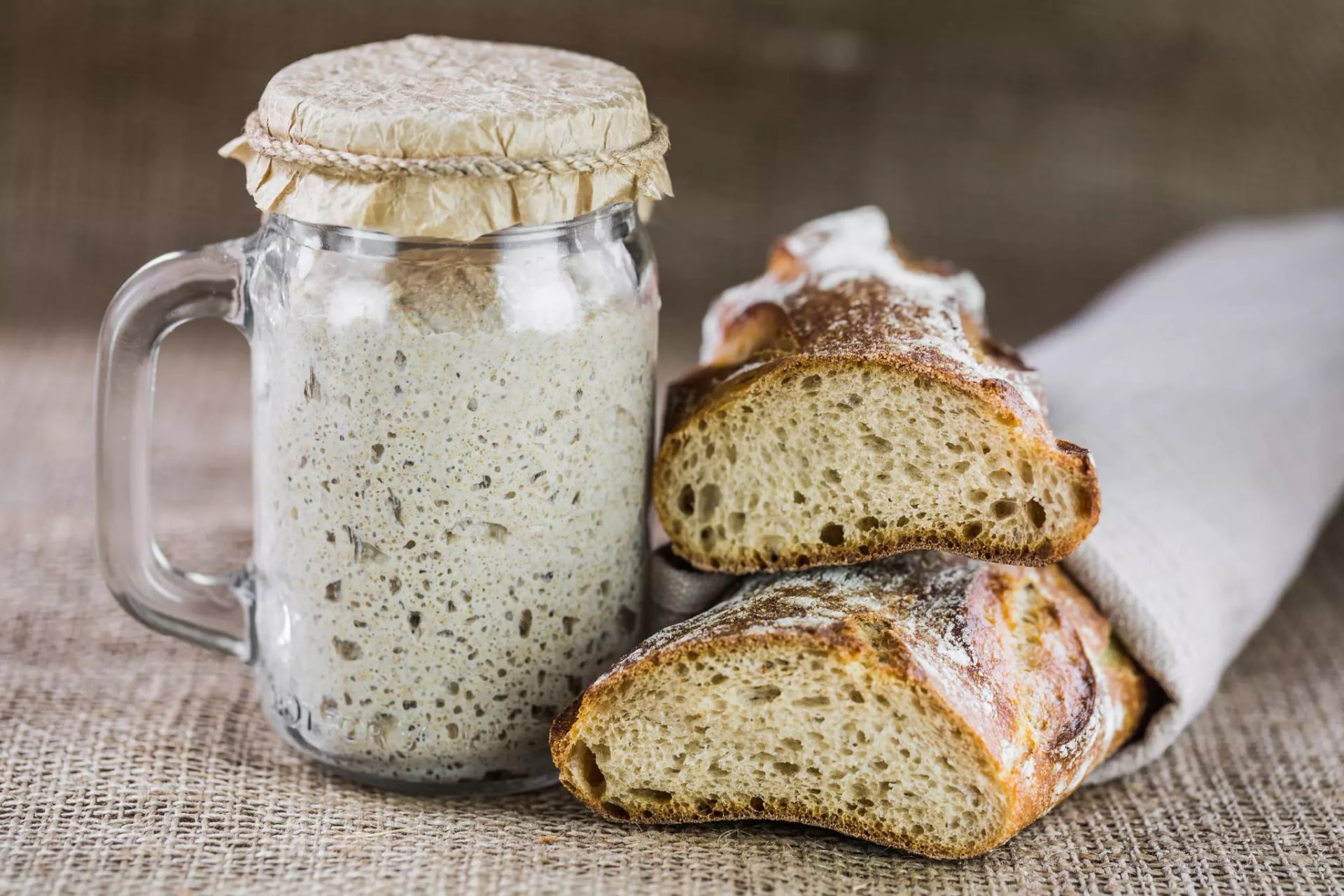 Ein Sauerteigstarter in einem Henkelglas, abgedeckt. Daneben frisches Baguette, aufgeschnitten.