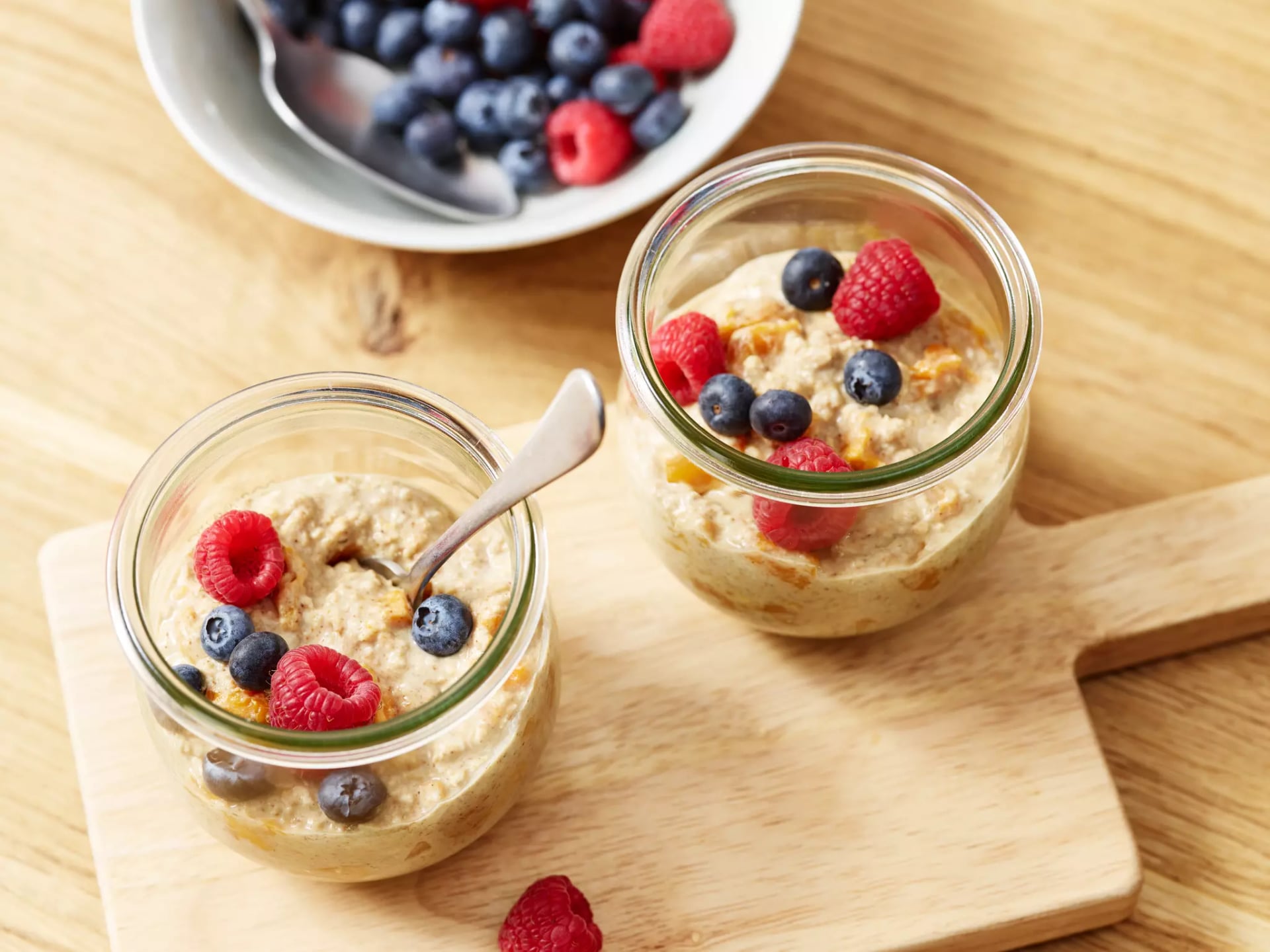 Overnight Oats mit Mango und Kokos abgefüllt in zwei kleinen Glässchen stehen auf einem Holzbrett.