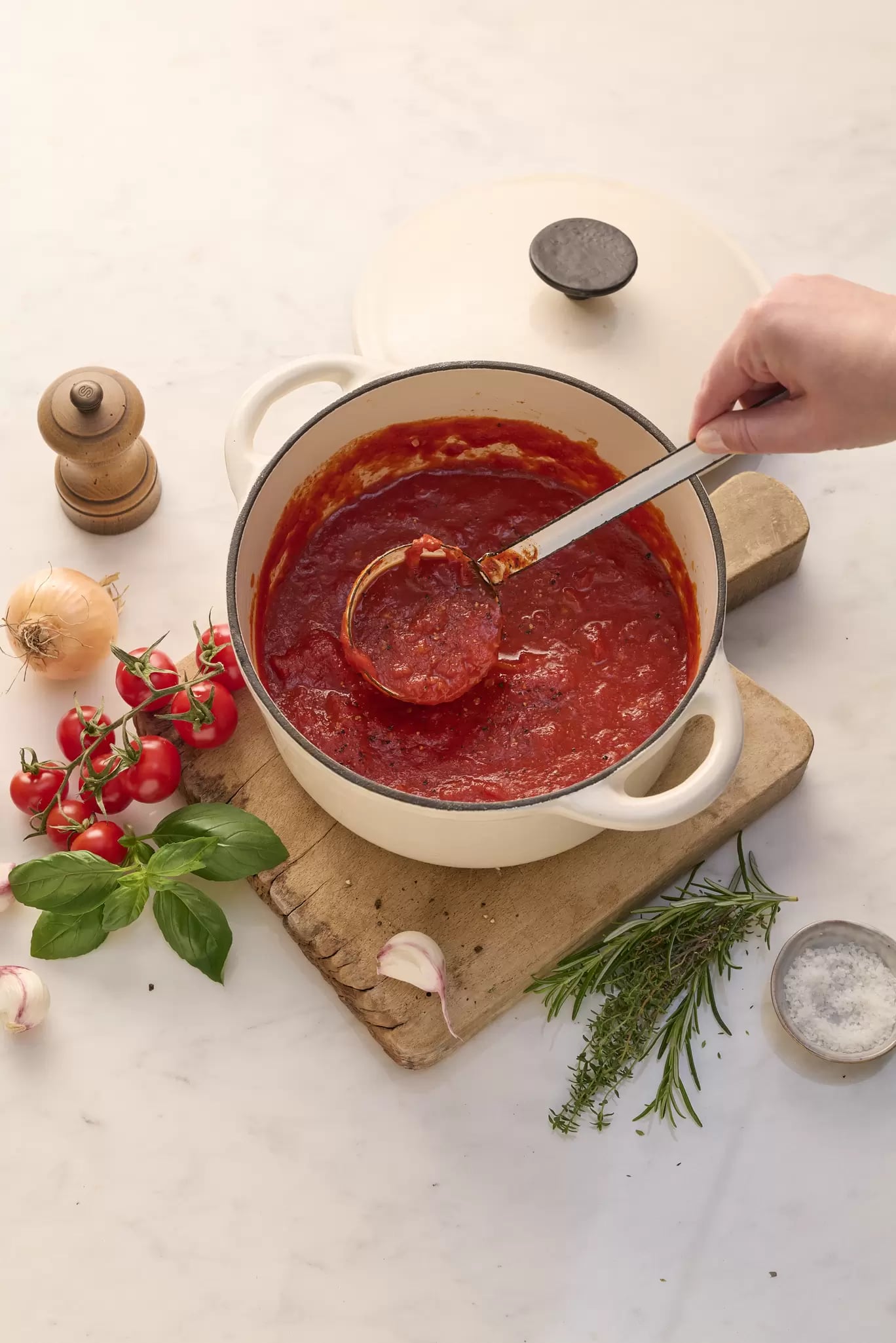 Das Bild zeigt eine weiße Schmorpfanne mit selbstgemachter Tomatensauce.  Eine Schöpfkelle mit Sauce wird herausgehoben.  Neben der Pfanne liegen auf einer Holzunterlage Cherrytomaten, Basilikum, Knoblauchzehen und eine Zwiebel. Im Hintergrund ist eine Pfeffermühle zu sehen. Das Gesamtbild wirkt appetitanregend und vermittelt ein Gefühl von hausgemachter, einfacher Küche.