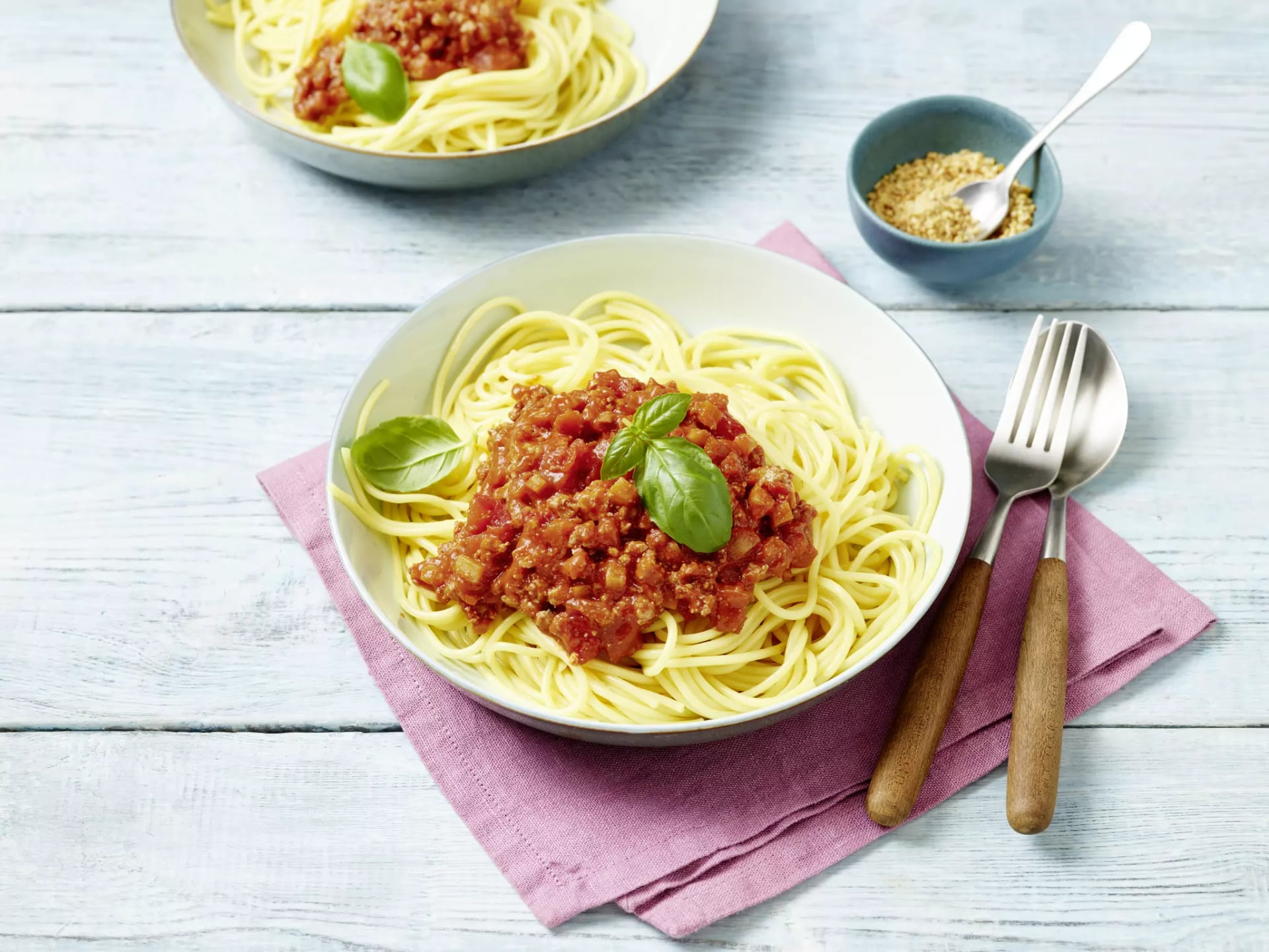 Teller mit Spaghetti und Tomatensoße, garniert mit Basilikum, daneben Besteck auf rosa Stoffserviette