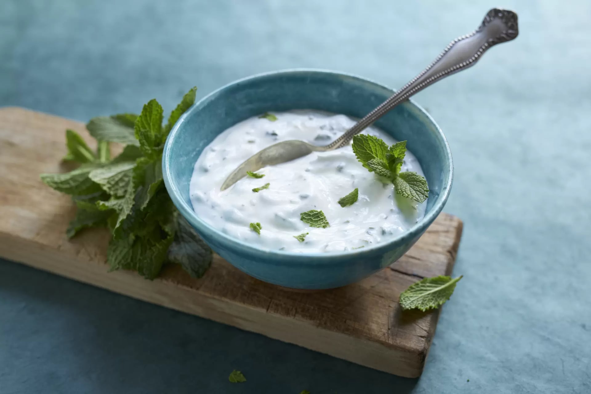 Blaue Schale mit weißem Joghurt und Minzblättern auf Holzbrett mit Silberlöffel