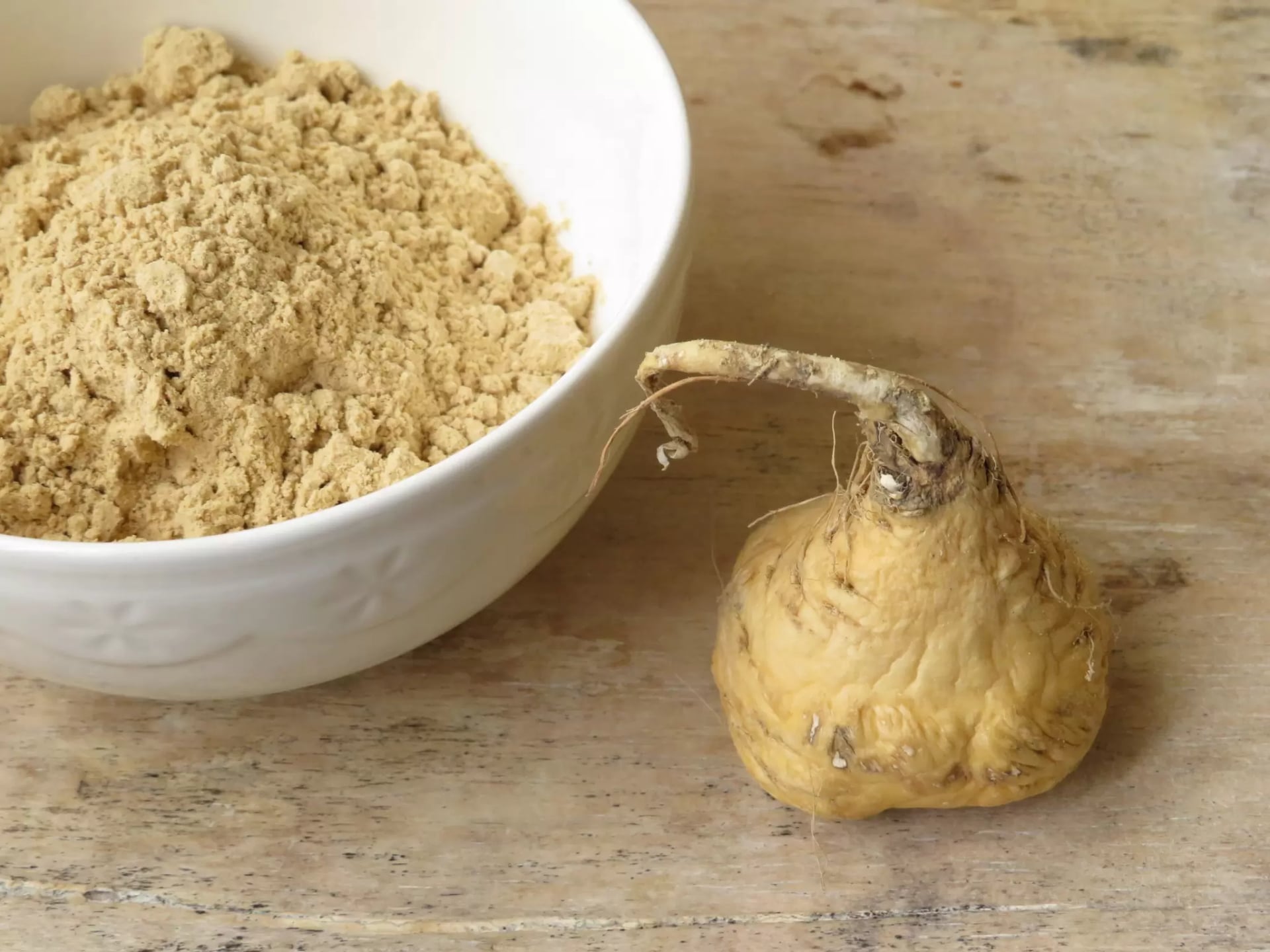 Eine Knolle frischer Maca steht neben einer weißen Schüssel gefüllt mit Maca Pulver