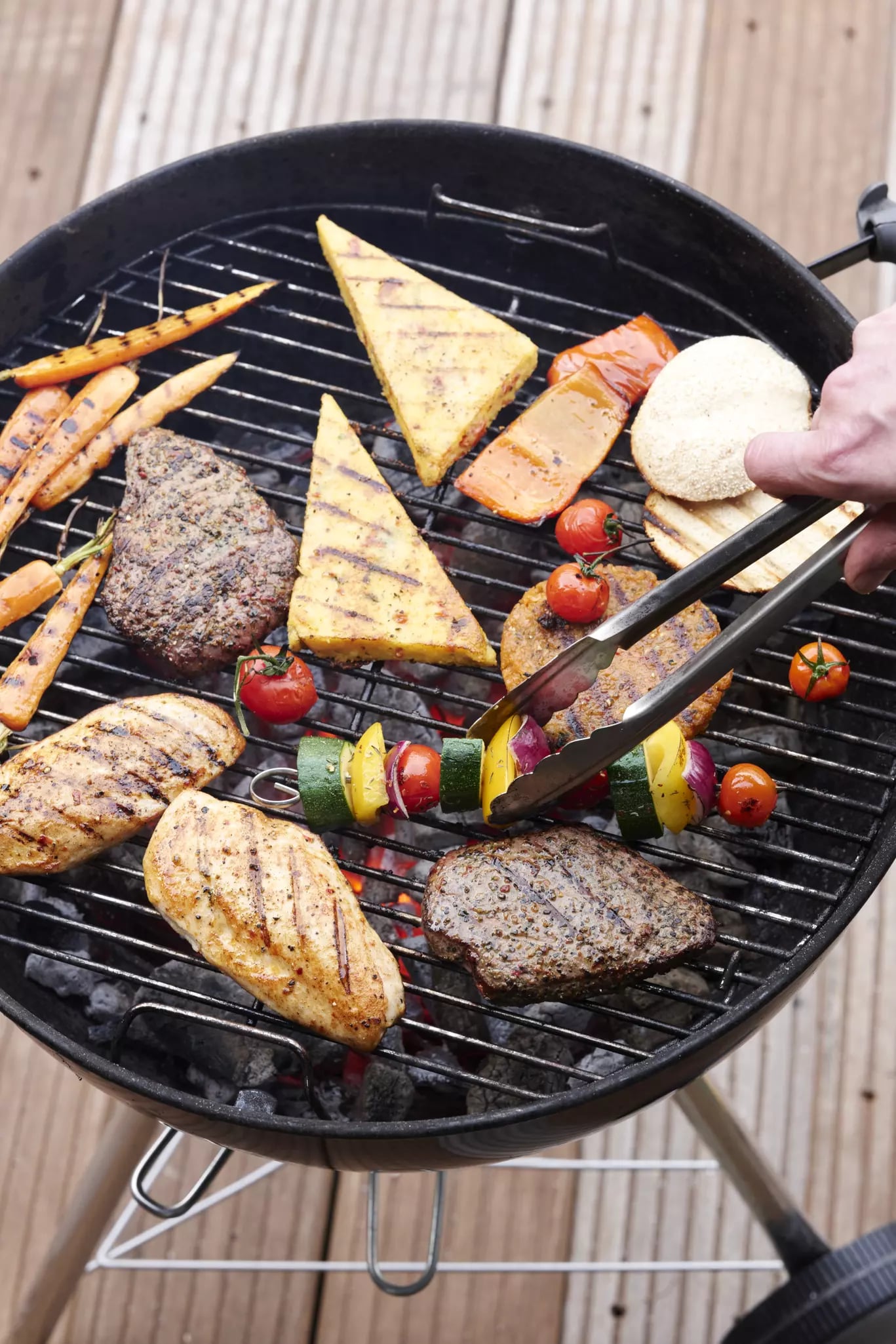 Auf dem Gitter eines Kugelgrills liegen verschiedene gegrillte Gemüse, Käse- und Fleischvarianten. Eine Person greift mit einer Grillzange nach einem Gemüsespieß.