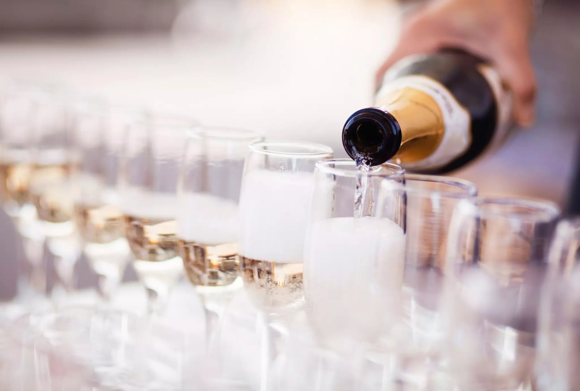 Eine Hand schenkt Schaumwein aus einer klassischen grün-goldenen Flasche in viele, aneinandergereihte Sektgläser ein