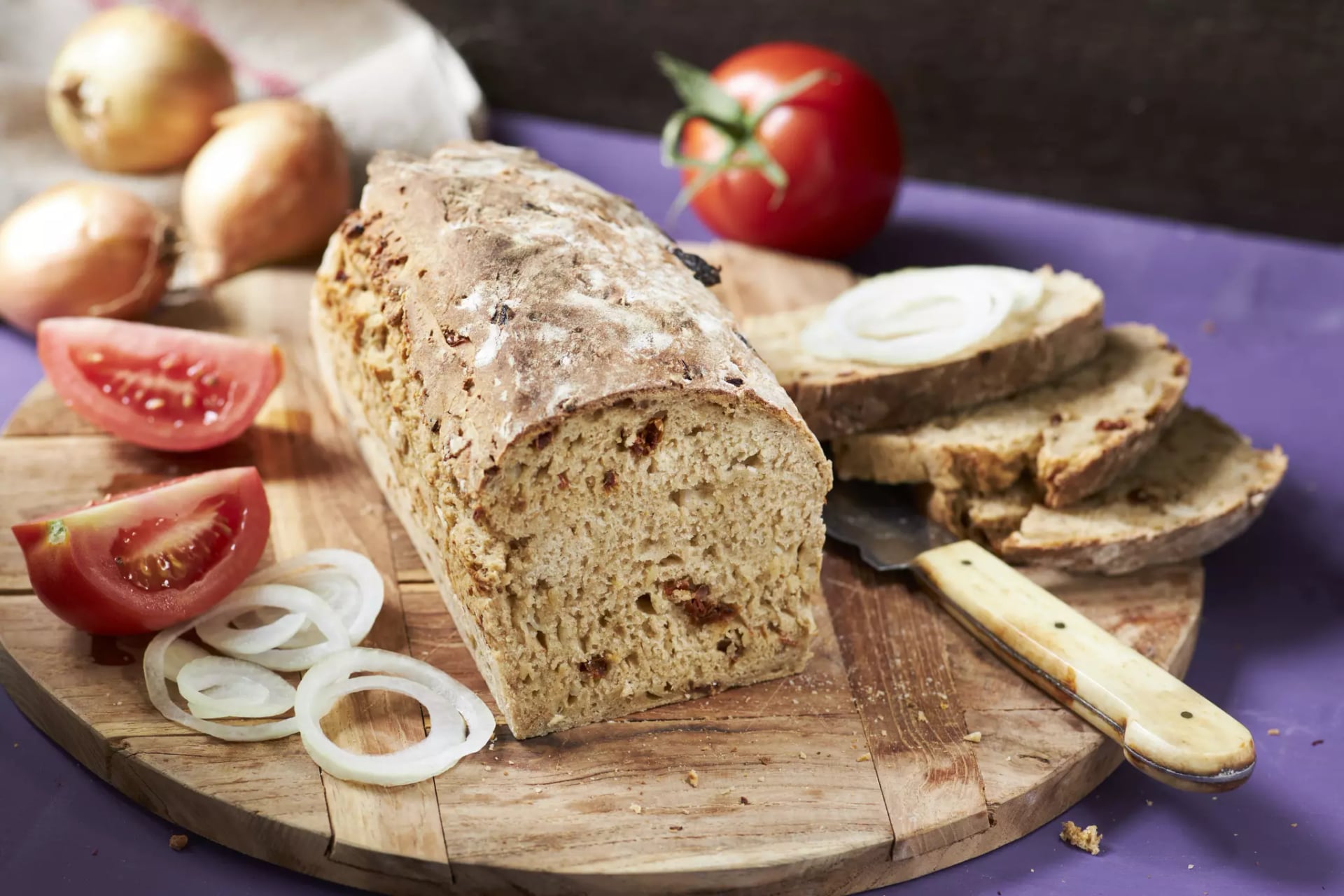 Laib Brot mit mehreren Scheiben, Tomatenvierteln, Zwiebelringen und Zwiebeln auf einem Holzbrett.