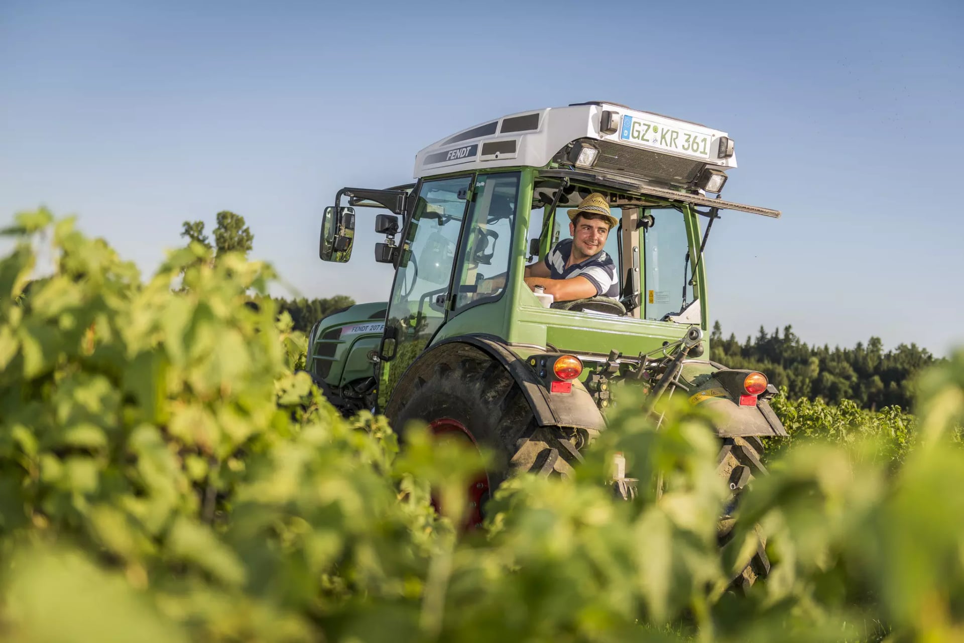Mann mit Hut sitzt lächelnd in grünem Traktor auf Feld mit Pflanzen.