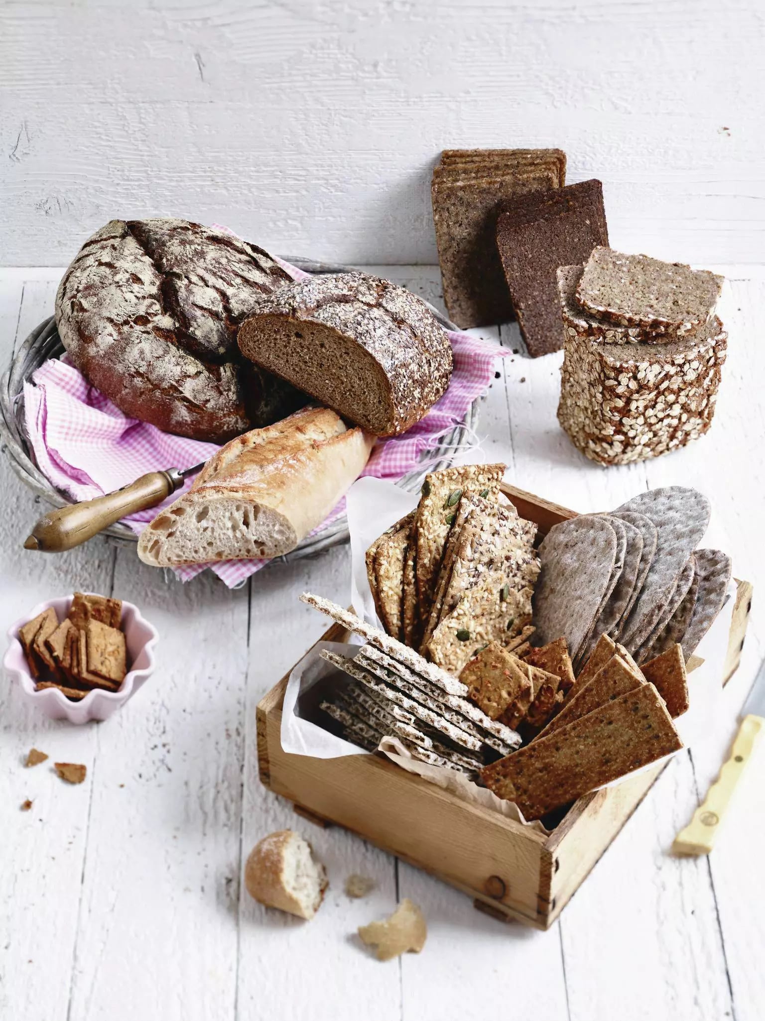 Verschiedene Brotsorten, darunter rundes Sauerteigbrot, Baguette, dunkles Vollkornbrot und verschiedene Knäckebrote in einer Holzkiste auf weißem Holztisch
